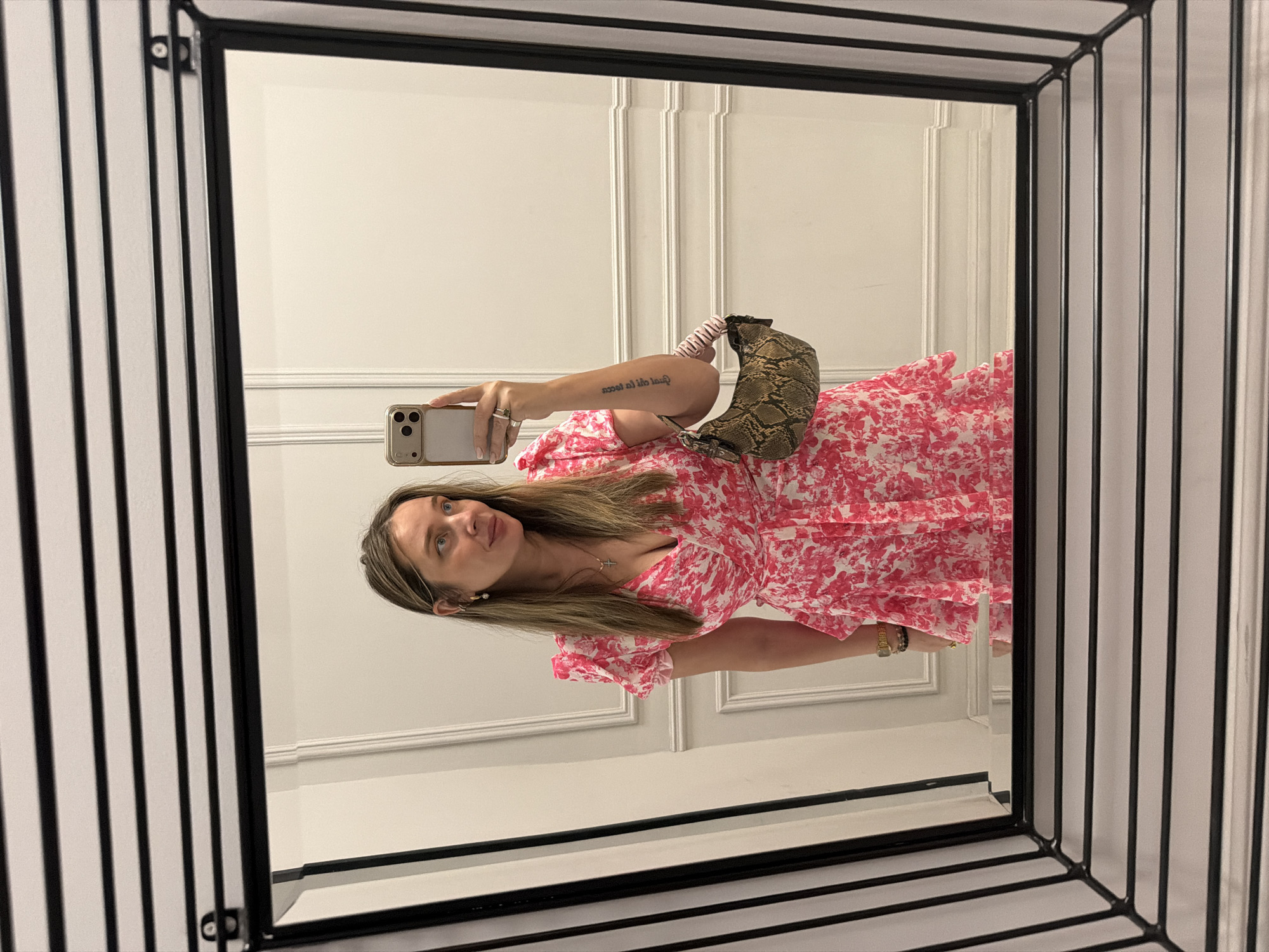 A woman is reflected in a mirror. She is holding a cell phone, taking a selfie. She has long brown hair, is wearing a floral pink and white dress, a gold necklace with a cross pendant, a watch, and gold earrings. She has a snake print handbag over her shoulder. The mirror has a black frame, and the wall behind her is white with decorative molding. The location cannot be determined from the image.