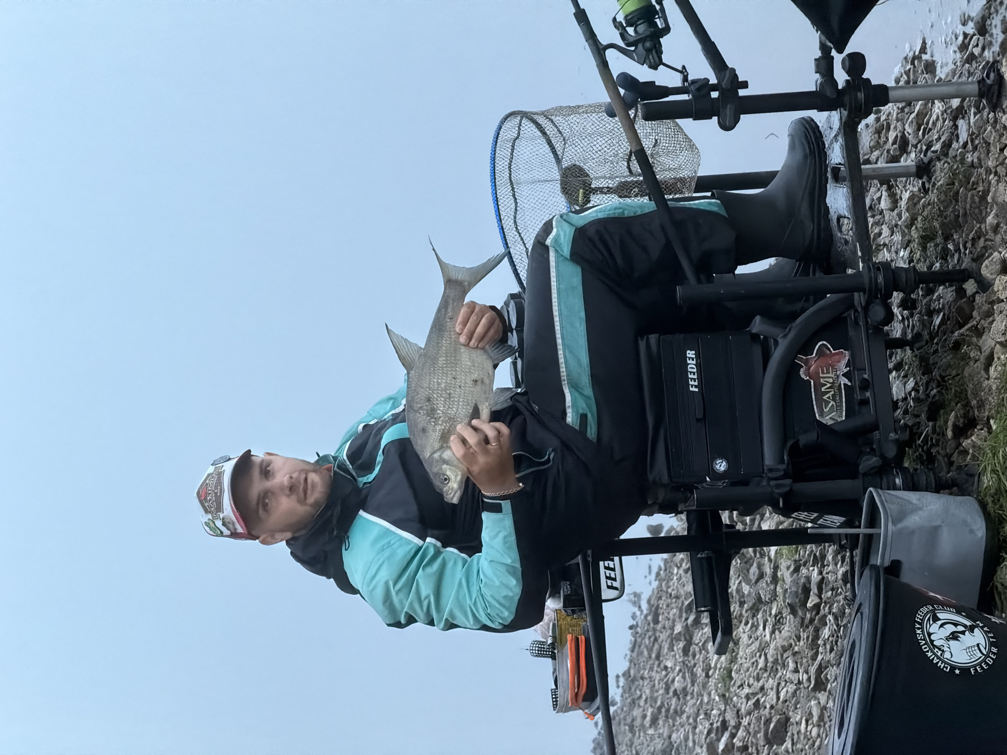 The image displays a man seated on a specialized fishing station on a rocky shore, holding a freshly caught fish. The man, seen from the waist up, wears a black and teal jacket, dark pants, and black rubber boots. He has a multi-colored cap on his head and looks towards the viewer with a calm expression. He holds a silver-scaled fish, likely a bream, horizontally with both hands, presenting his catch.
Behind the man, fishing rods are set up in holders, and a landing net is visible. The fishing station itself is robust, with various compartments and attachments. The ground is covered with light-colored rocks and pebbles, extending to the edge of a body of water visible on the right side of the frame. The sky, which occupies a large portion of the background, is a uniform light blue-grey, indicating an overcast day with soft, diffused lighting.
Visible text includes "FEEDER" on the side of the fishing station, "ASAME" on an attached bag, and a black bucket in the foreground proudly displays a white skull logo with "CHAIKOVSKY FEEDER CLUB TEAM". Text on the man's cap includes "Marmara". The scene captures a moment of successful fishing in Noviy, Russia, under an overcast sky.
