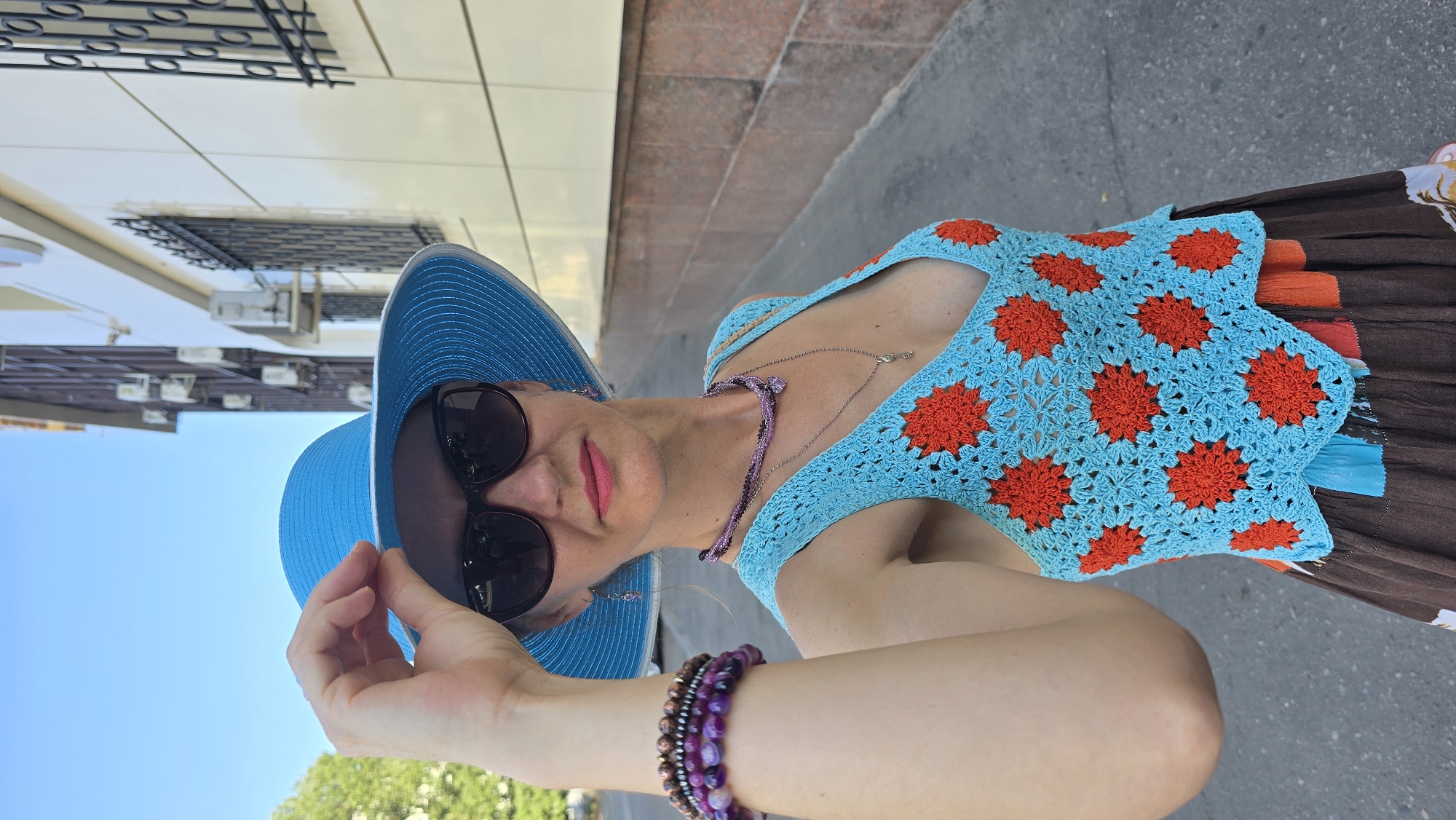 A woman in a blue sun hat and a crocheted turquoise top with orange floral patterns stands on a sidewalk. She is wearing sunglasses and jewelry. A building with multiple windows and air conditioning units is visible behind her, along with trees and a blue sky. The image appears to have been taken in a city environment.