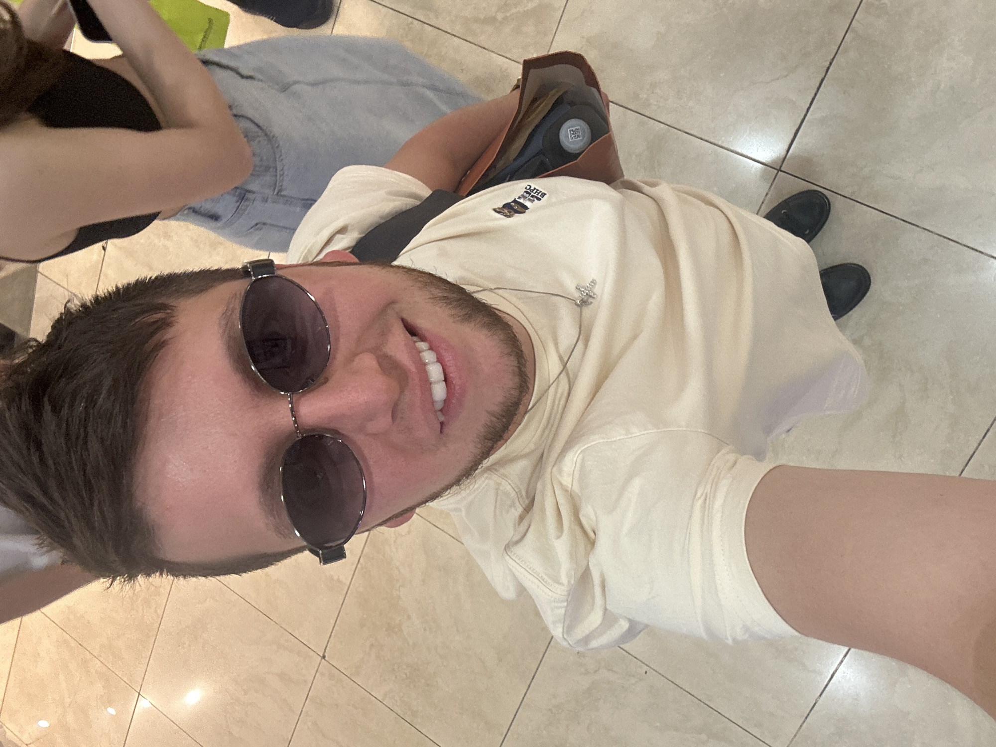 A high-angle, eye-level, full-body selfie of a Caucasian male in his early 20s is shown, standing indoors on a tiled floor. He has light brown hair, a short beard, and is wearing sunglasses, a light tan t-shirt with a small graphic on the left chest, a silver cross necklace, and black loafers. He is smiling, with his mouth open. To his right, a person's legs are visible, wearing a black tank top and light blue jeans. In the male subject's right hand, he is holding a brown bag that contains a dark bottle. The background is out of focus, but appears to be a public indoor space with tiled flooring and possibly a store. There is no indication of the location being Moscow, Russia.