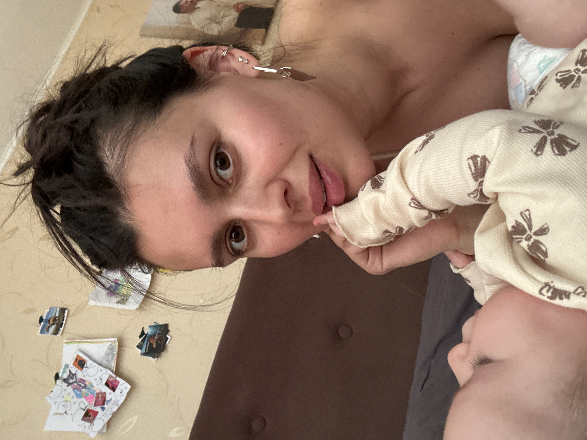 A woman with dark hair pulled back in a bun is lying on her side, looking at the camera. She has fair skin, brown eyes, and is wearing a silver earring. Her mouth is slightly open. She is holding a baby in her arms. The baby is wearing a cream-colored onesie with brown flower patterns. The baby's face is partially visible. The background is a light yellow wall with small items attached. A portion of a headboard is visible behind the woman. The location is Chaikovsky, Russia.