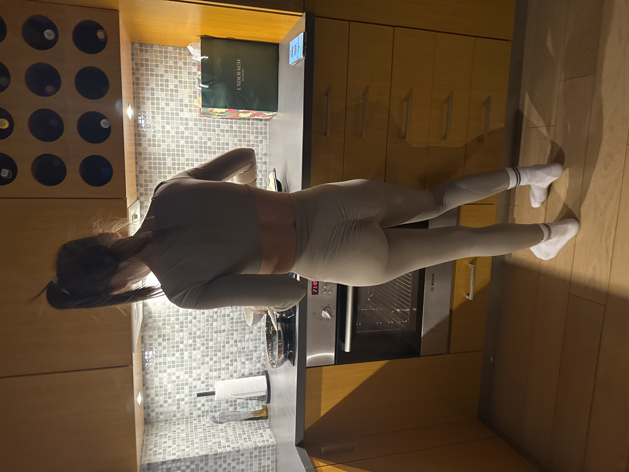 The image captures a modern kitchen scene in Warsaw, Poland, during the evening, indicated by the oven's digital display showing "21:10." A person, seen from behind, stands at a dark grey countertop. They have dark hair pulled back in a ponytail and are dressed in light beige athletic wear, including a long-sleeved top and matching leggings, finished with white socks featuring black stripes.
The kitchen is equipped with light wooden cabinetry and a backsplash made of small, light-colored square tiles. Under-cabinet lights illuminate the workspace. On the countertop, a metal pan or bowl, a paper towel roll on a holder, and a small electronic device are visible. A green shopping bag with "LADERACH" printed on it rests nearby. Below the counter, a "BOSCH" brand oven is integrated. To the left, a wooden wine rack holds several bottles. The flooring is composed of light wooden planks. The person appears to be engaged in an activity at the counter, possibly food preparation.
