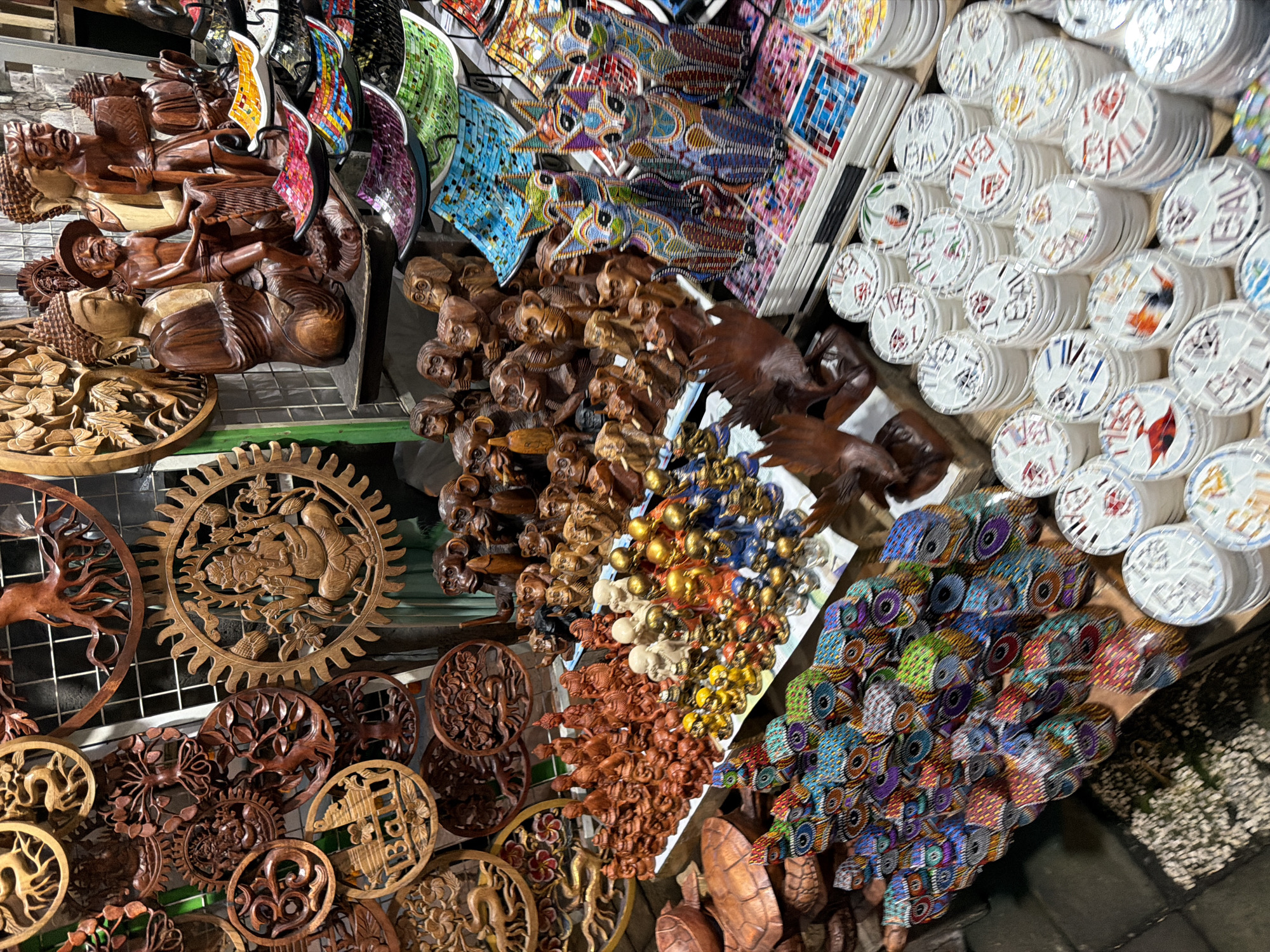 This is a detailed, eye-level, medium shot of a market stall in Ubud District, Indonesia, displaying a wide variety of handcrafted items. The stall is densely packed with wooden carvings, including intricate circular pieces with motifs like trees, a deer, and a depiction of Ganesh. There are also numerous smaller wooden figures, such as monkeys and Buddhas, in various poses and sizes. In the foreground, stacks of colorful, mosaic-covered dishes are visible, as well as decorative mosaic cats and owls. Further back, a collection of stacked circular coasters, many with "BALI" written on them and mosaic designs, are arranged. The lighting is moderate, with some shadows, suggesting an indoor or covered outdoor market setting. No people are directly visible in the image, but the arrangement implies an active marketplace.