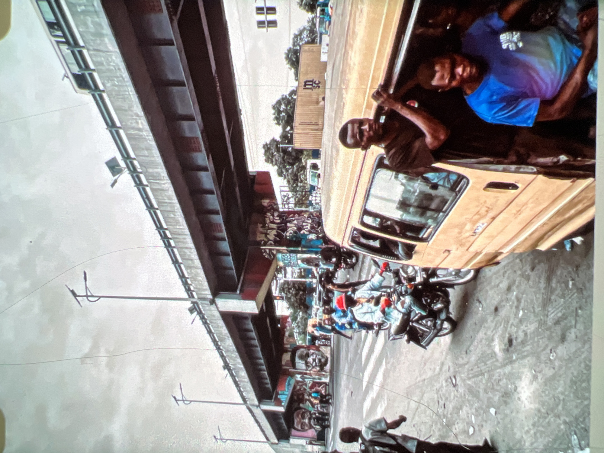 The image depicts a street scene in Lagos, Nigeria, under an elevated highway. In the foreground, a yellow commercial van is partially visible on the right. Two men are near the open passenger door of the van; one, wearing a dark t-shirt, is holding onto the roof of the van, while another, in a blue t-shirt, is seated inside. Further down the street, a man in a red hat is riding a motorcycle, with another person behind him. Several other motorcycles and pedestrians are visible, suggesting busy street life. The elevated highway stretches across the upper portion of the image, supported by concrete pillars. Graffiti art adorns the pillars and walls beneath the highway. In the background, trees and buildings are visible, along with shipping containers. The sky is overcast, and the ground is a paved road with some debris.