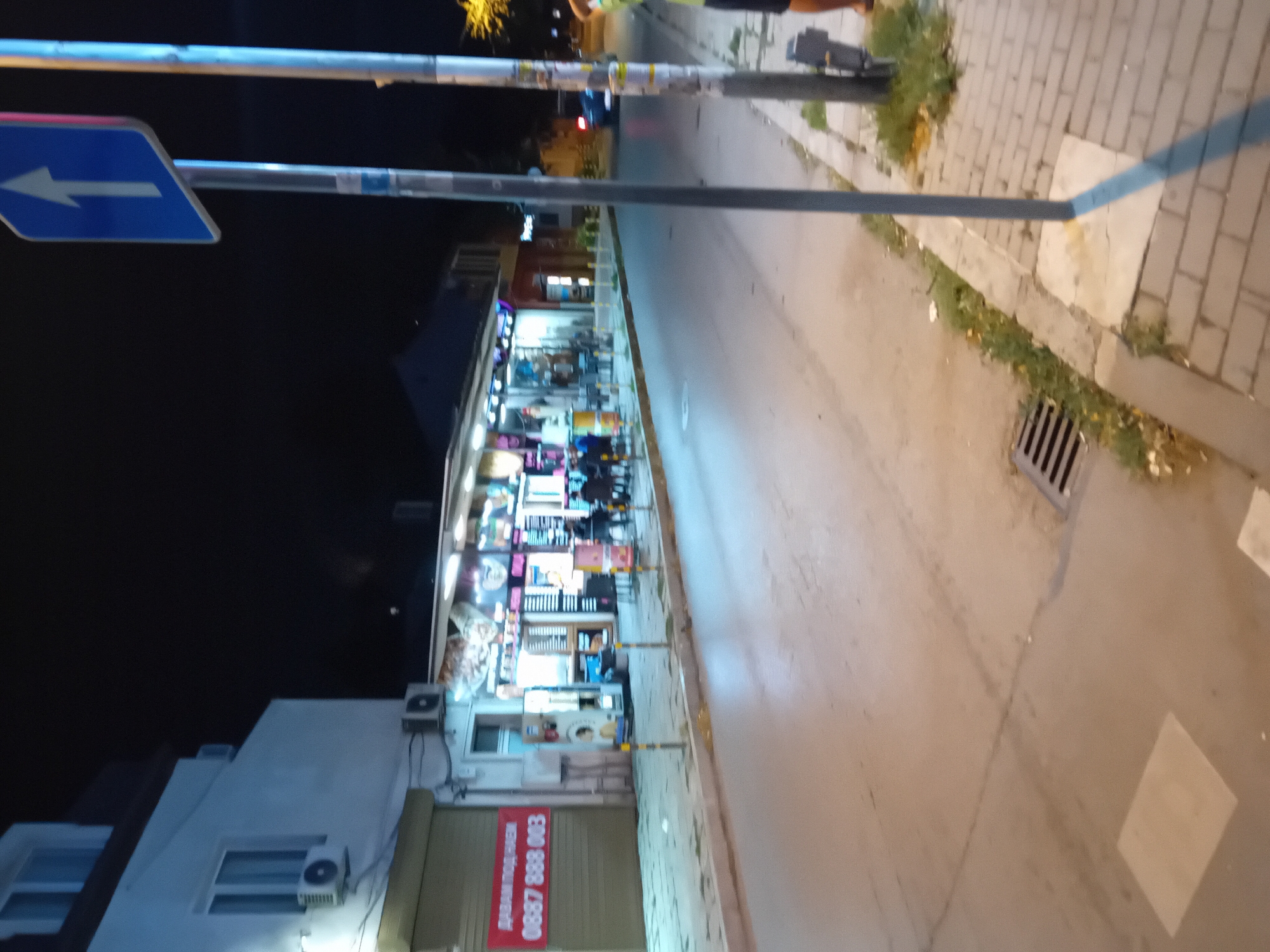The image captures a nighttime street scene in Varna, Bulgaria. A white building with multiple windows and an air conditioning unit is visible on the left, with a red banner displaying text and a phone number. Adjacent to it, a row of illuminated businesses lines the street, with several tables and chairs outside some establishments. People are seated at some of the tables.
A prominent blue traffic sign with a white arrow pointing upwards stands on the right side of the street, attached to a metal pole. Another pole with signage is situated next to it. The street itself is paved, with a sidewalk constructed of brick pavers. A storm drain is visible in the street. The overall lighting suggests it is nighttime, with artificial lights from the businesses and streetlights illuminating the scene. A portion of a blue car and the lower body of a person are visible on the far right edge of the image.