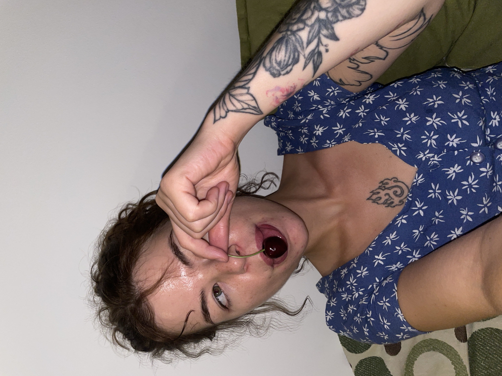 A young woman with curly brown hair and freckles is holding a cherry near her mouth, with her tongue slightly extended. She is wearing a blue dress with a white floral pattern and has tattoos on her arm and chest. The background is a plain white wall. The scene appears to be indoors, possibly in a home setting, and does not provide any specific visual cues to indicate the city of Boreti, Montenegro.