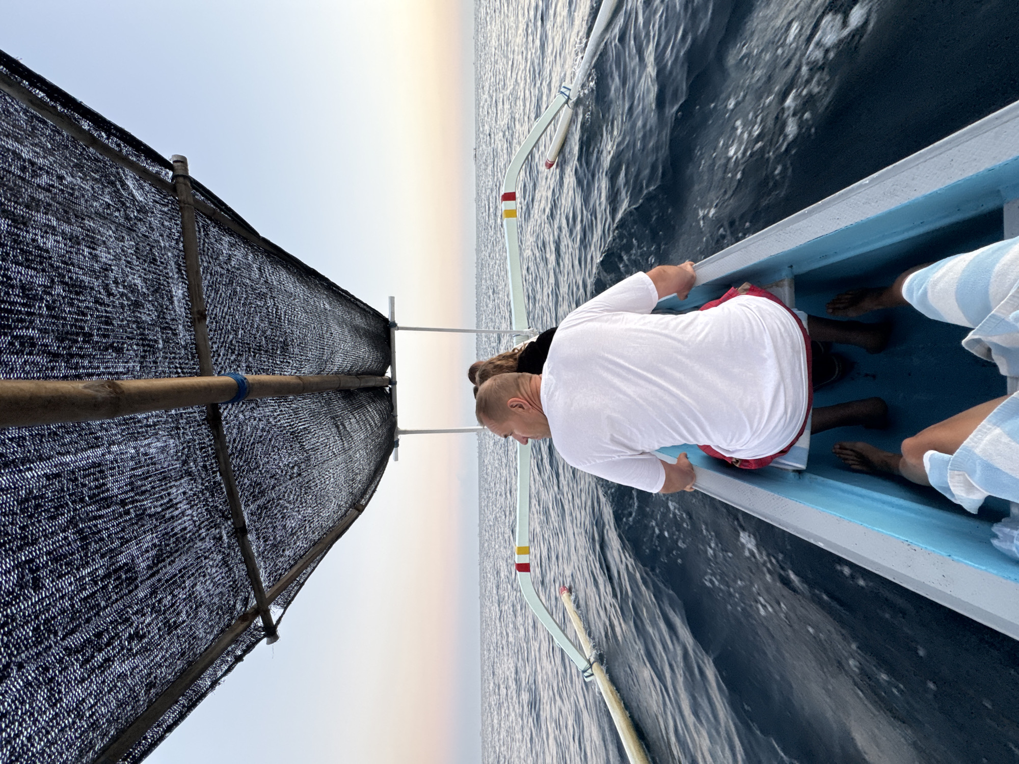 A medium shot captures a man in a white t-shirt and red shorts sitting in a traditional wooden boat, facing away from the camera and looking out at the ocean. The boat, with its blue interior and white-rimmed edges, is moving through dark blue water, creating a wake. Overhead, a black mesh tarp supported by bamboo poles provides shade. Two other individuals are seated further back in the boat, their presence indicated by their backs and hair. The background consists of the calm ocean meeting a pale pink and blue sky, suggesting either sunrise or sunset. The location is identified as the waters near an unknown city in Indonesia.