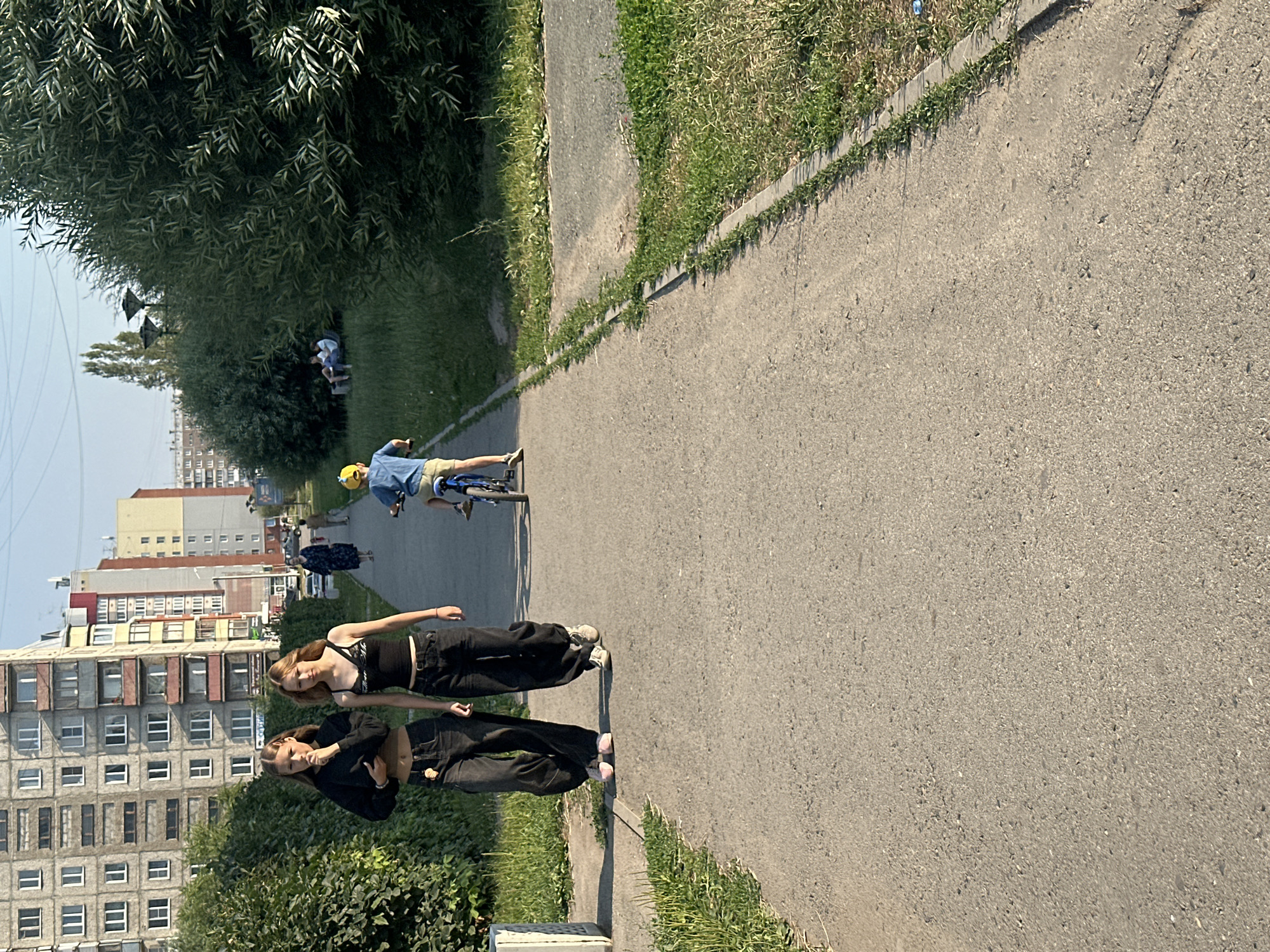 The image depicts a paved pathway in a residential area of Novoaltaysk, Russia. Two young women, dressed in casual dark clothing, are walking towards the camera on the left side of the path. To the right of the path, a young child wearing a yellow hat and blue shirt is riding a bicycle away from the camera. In the background, there are apartment buildings, trees, and a grassy area where a few people are seated on a bench. The scene appears to be set during daylight hours, with sunlight casting shadows on the ground.