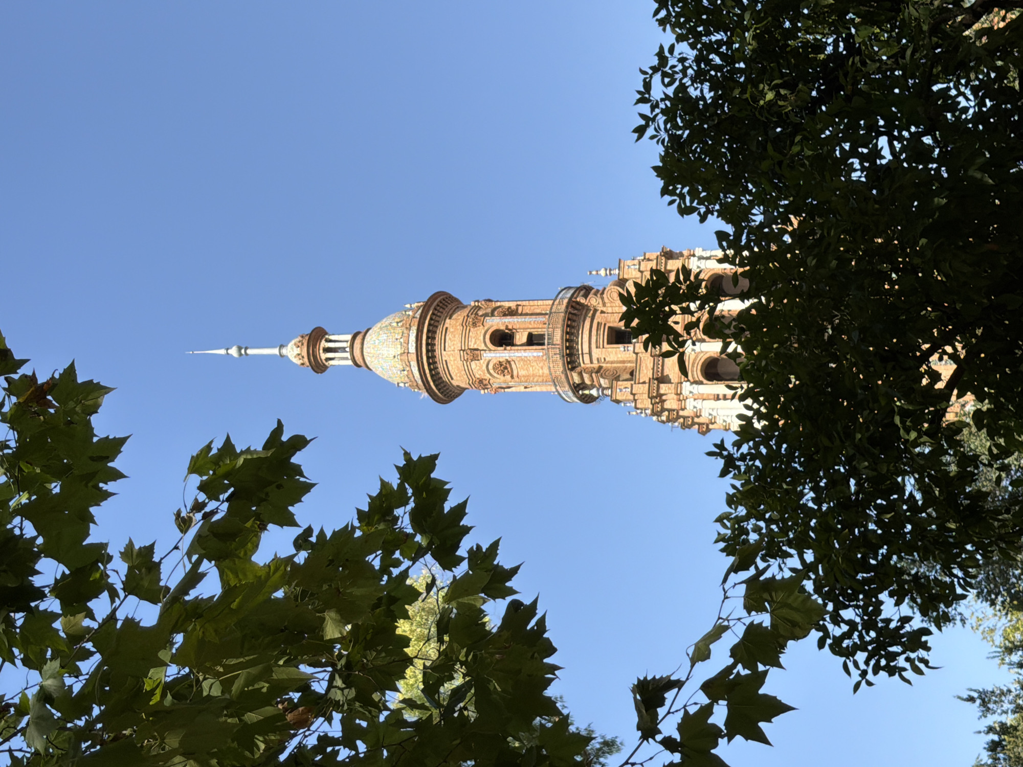 The image depicts the Torre de San Vicente, a Gothic tower in Seville, Spain, framed by lush green foliage. The tower, constructed of brick, features ornate architectural details, including arched windows and decorative elements around its uppermost sections. A slender, pointed spire crowns the tower, reaching towards a clear, bright blue sky. The foliage in the foreground partially obscures the tower, creating a sense of depth and framing the architectural subject. The scene is captured in daylight, suggesting a pleasant, sunny day in the city.