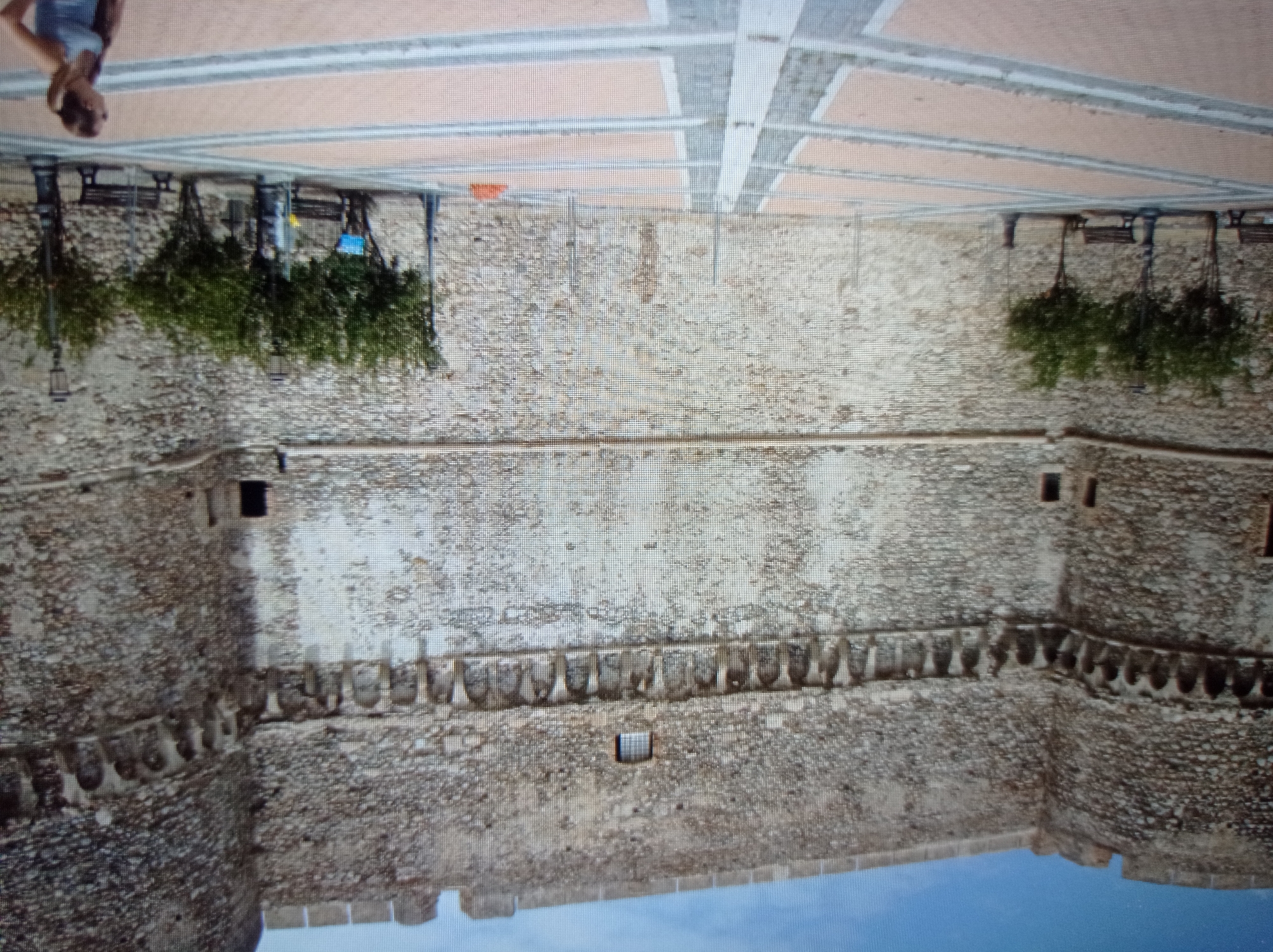 The image depicts the exterior of a large, stone fortress. The fortress walls are constructed of rough-hewn stones and feature a crenellated parapet along the top. Several arched openings are visible along a horizontal band just below the parapet. A small, rectangular window is situated higher up on the wall.
In the foreground, a paved courtyard with a patterned design of alternating light and darker rectangular sections leads up to the fortress. Several benches and street lamps are placed in the courtyard. Small trees are planted in front of the fortress wall.
A woman with brown hair tied in a ponytail is visible in the lower right portion of the image, looking to the left. She is wearing a light-colored tank top and has her right hand on her hip.
The image does not provide any information that places it in the city of Mukachevo, Ukraine.