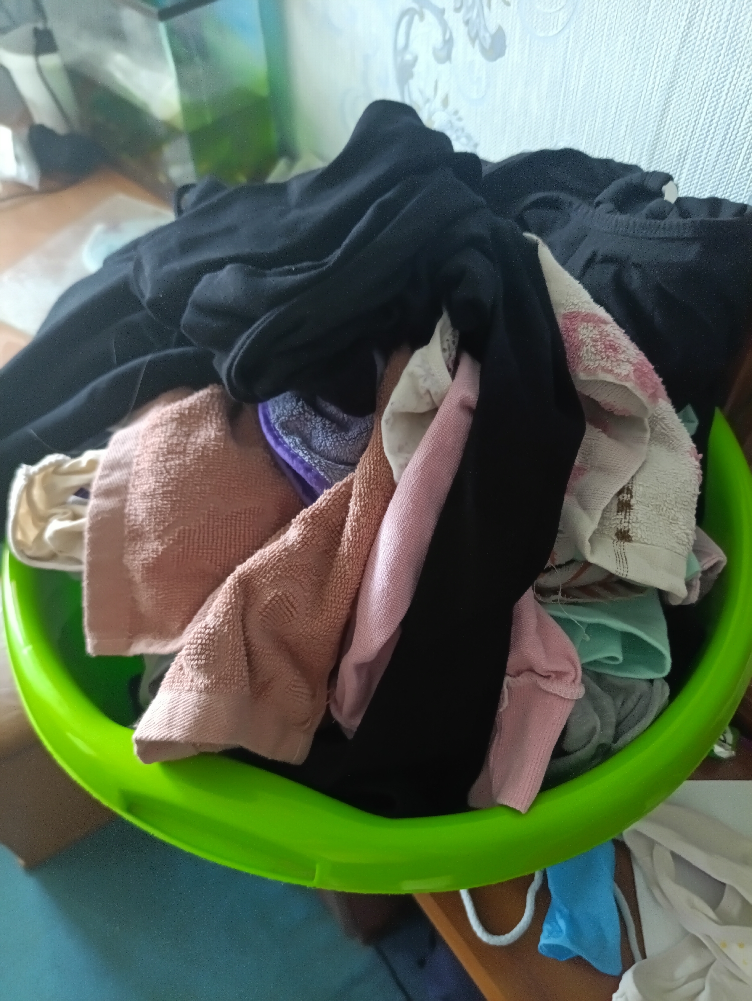 A vibrant green plastic laundry basket is overflowing with various articles of clothing. The laundry is a mix of colors and textures, including dark black garments, pale pink fabric, and beige and peach-colored towels with textured patterns. A white towel with a pink floral design is also visible. Some of the clothing appears to be folded or bunched up, while other pieces are draped over the sides of the basket. The scene appears to be indoors, with a textured wallpaper visible in the background. A wooden surface and a hint of a blue floor are also present. The lighting suggests daytime. The setting is likely a home environment in Kamianets-Podilskyi, Ukraine, where laundry is being prepared or has been recently collected. There is no visible text in the image.