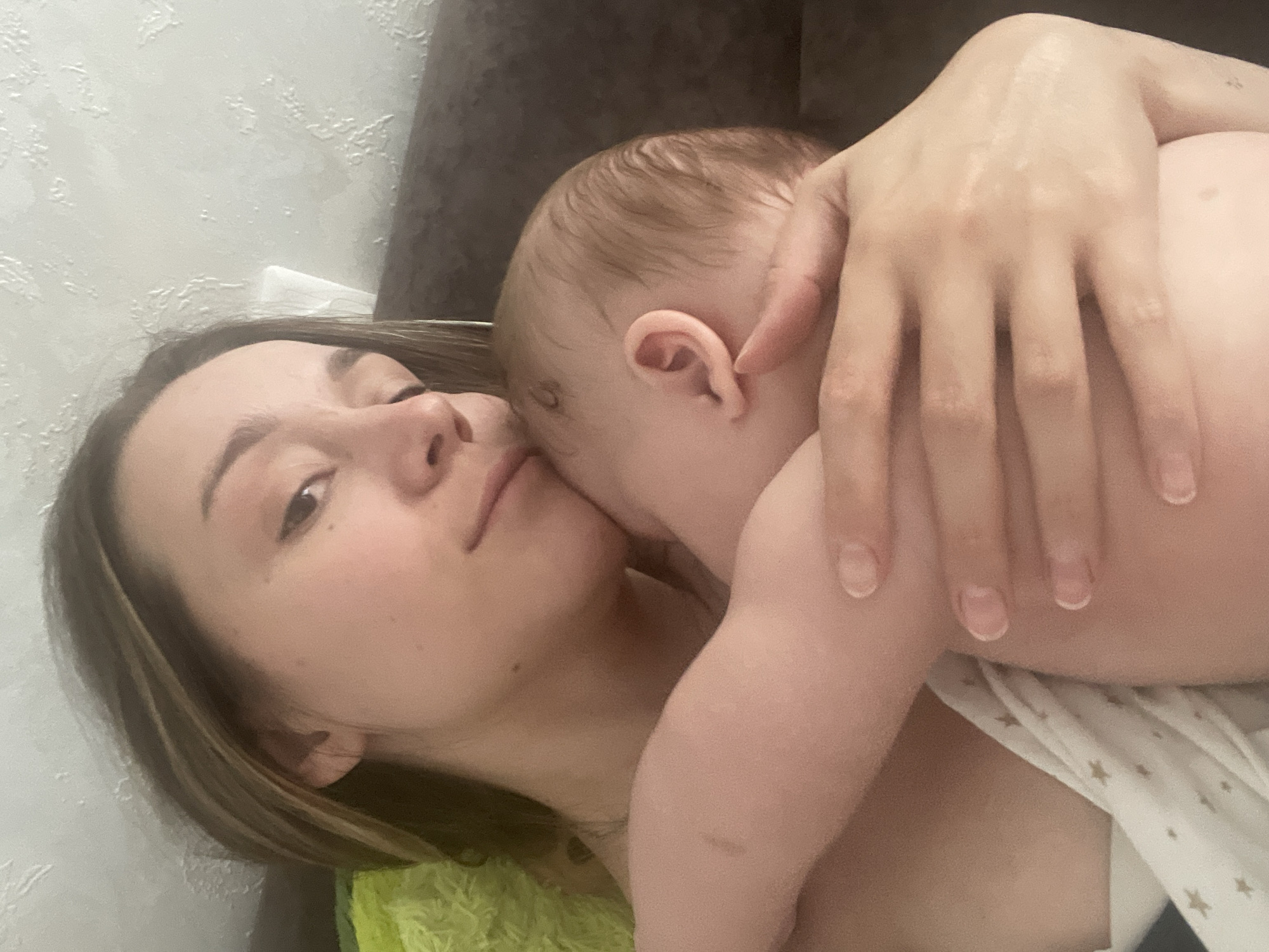 The image is a close-up, indoor photograph of a woman holding a sleeping baby. The woman's head is tilted down, resting against the baby's head, and her arms are wrapped around the infant. The baby is facing away from the camera, with its head nestled against the woman's shoulder. The woman has long, light brown hair that is partially covering her face. The baby has light brown hair and appears to be very young. They are both wearing casual clothing. The background is blurred, suggesting a domestic setting. There is no indication of the city of Stavropol, Russia, or any other specific geographical location. The image appears to be taken with a smartphone or similar device, as evidenced by the lighting and composition.