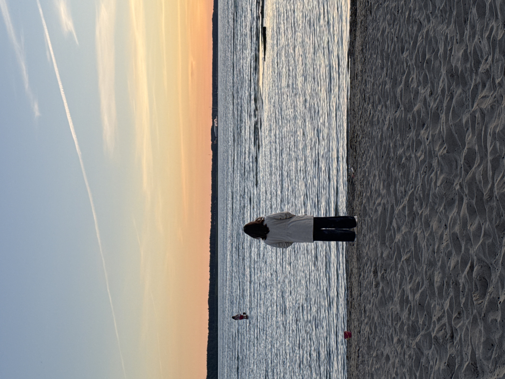 An outdoor, medium shot captures a sunset over a body of water, with a sandy beach in the foreground. A lone person stands facing the water, their back to the camera, wearing a light-colored jacket and dark pants. In the distance, another person is visible in the water, along with a red bucket on the sand near the primary subject. The sky displays a gradient of orange and blue with a faint contrail. The scene appears to be from a coastal area in Tallinn, Estonia.