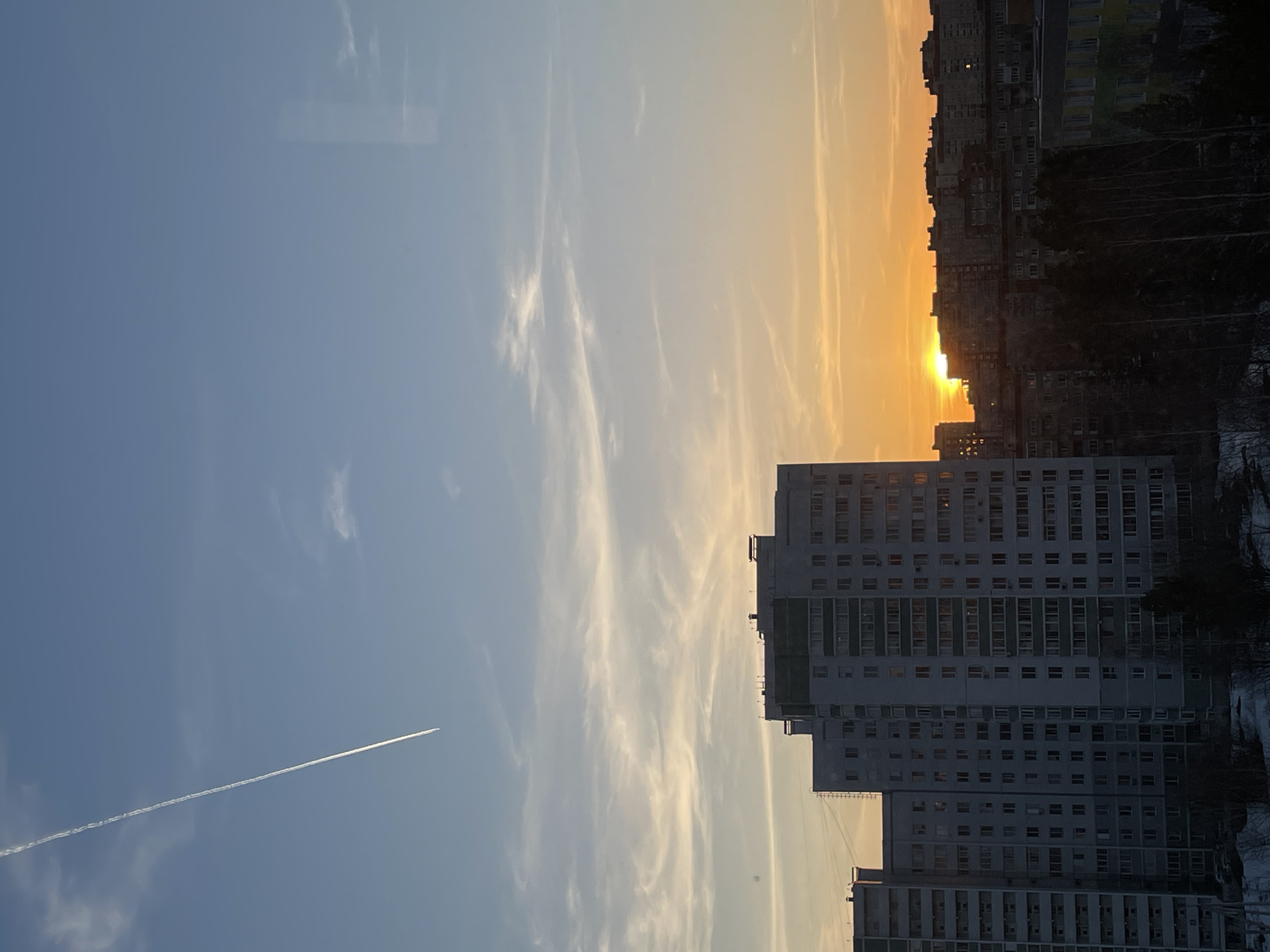 The image presents a cityscape, likely taken at either dawn or dusk, due to the presence of a brightly lit sun near the horizon and the varying shades of light in the sky. The scene is dominated by a large, multi-story apartment building, possibly made of concrete or similar material. Its facade is punctuated by numerous windows, implying high-density residential occupancy. Behind this building, several other structures are visible. The presence of bare trees suggests it might be late autumn or winter.
The sky features a gradient of colors, from a deep blue higher up to a warm, orange-yellow near the horizon, where the sun appears to be setting. Wisps of clouds are present, some catching the light of the setting sun. A single contrail from a high-altitude aircraft cuts across the blue expanse, adding a sense of scale to the scene.
Determining the location definitively as Izhevsk, Russia, requires additional information like specific architectural details, local landmarks visible in the wider panorama, or specific identifying details associated with the city. Without such details, pinpointing the exact location is impossible.