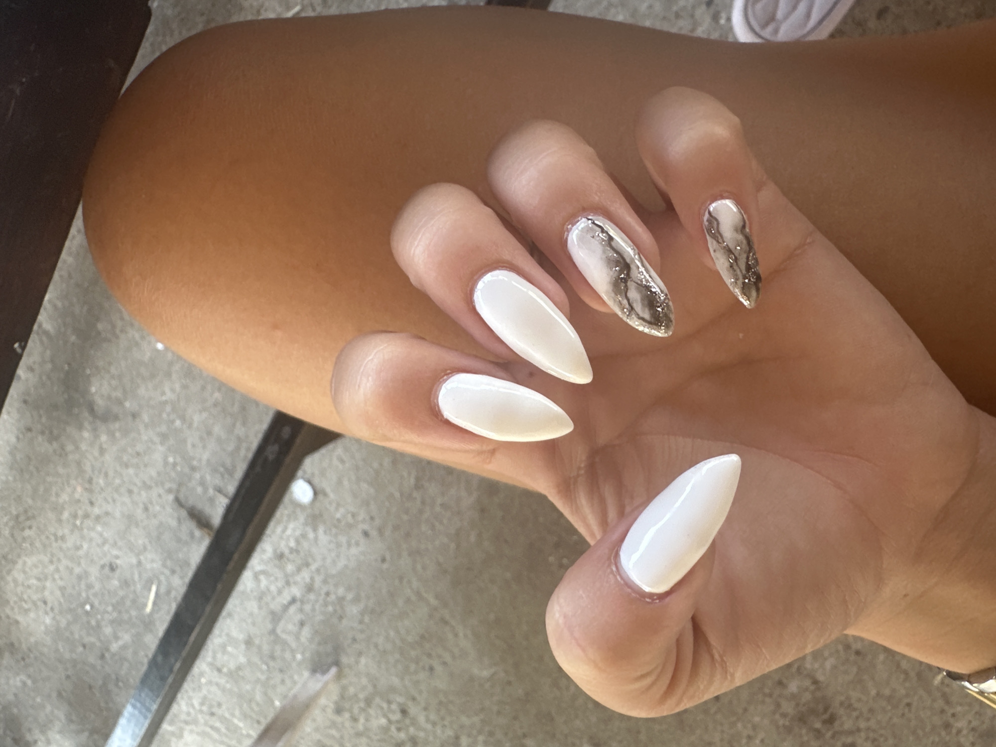 A person's hand with manicured nails is held in front of their leg and a concrete surface. The nails are stiletto-shaped. Four nails on the visible hand are white, and two nails have a marble design with white, gray, and black swirls, with some glitter accents. The person is wearing a watch with a silver band on their left wrist. A dark, metal object is visible in the background, as is a portion of a white shoe. The scene appears to be outdoors, possibly in an urban setting in Galiche, Bulgaria.