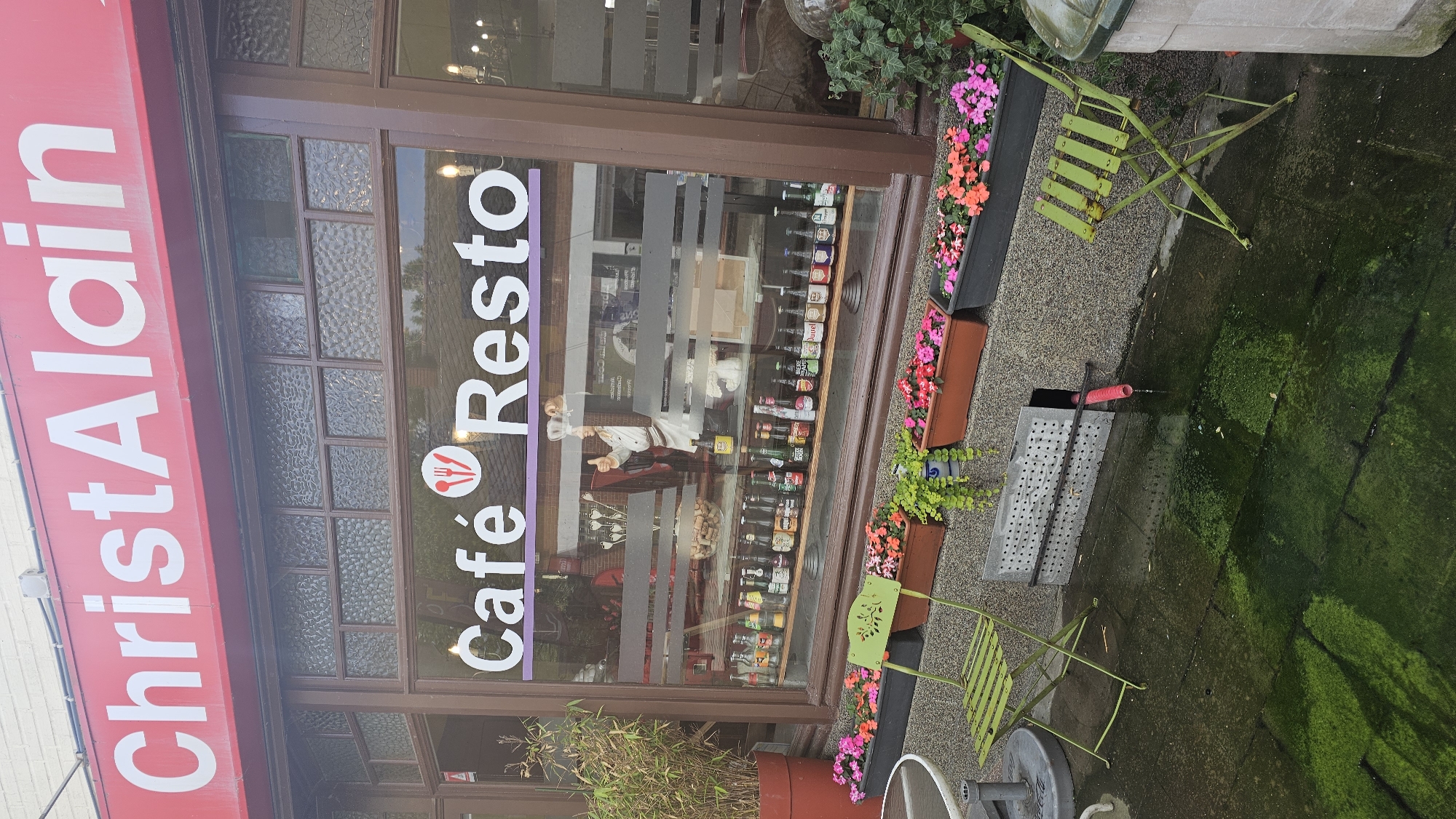 This image captures a street-level view of the exterior of a cafe or restaurant named "ChristAlain Café Resto" in Thuin, Belgium. The scene appears to be set during the daytime, with natural light illuminating the facade.
The building features a prominent red awning at the top with "ChristAlain" written in large white capital letters. Below the awning, there's a large display window with brown frames, part of which has textured glass panels. Across the main window, "Café Resto" is displayed in white text on a purple horizontal stripe, accompanied by a small icon of a fork and knife.
Through the window, various bottles are visible, neatly arranged on shelves inside the establishment. A cutout or poster of a chef figure, dressed in white, is also prominently displayed inside, giving a thumbs-up gesture.
Outside the cafe, there's a small seating area. Two light-green metal folding chairs are placed on the pavement. Several flower boxes filled with vibrant pink and orange flowers add a splash of color. A large red planter with bamboo-like plants is visible on the far left. The ground is paved but appears damp, with noticeable patches of green moss or algae, especially in the foreground and towards the right, suggesting a moist environment or recent rain. A metal grate and a red pipe are also present on the ground near one of the chairs. No direct human activity is depicted in the static scene.