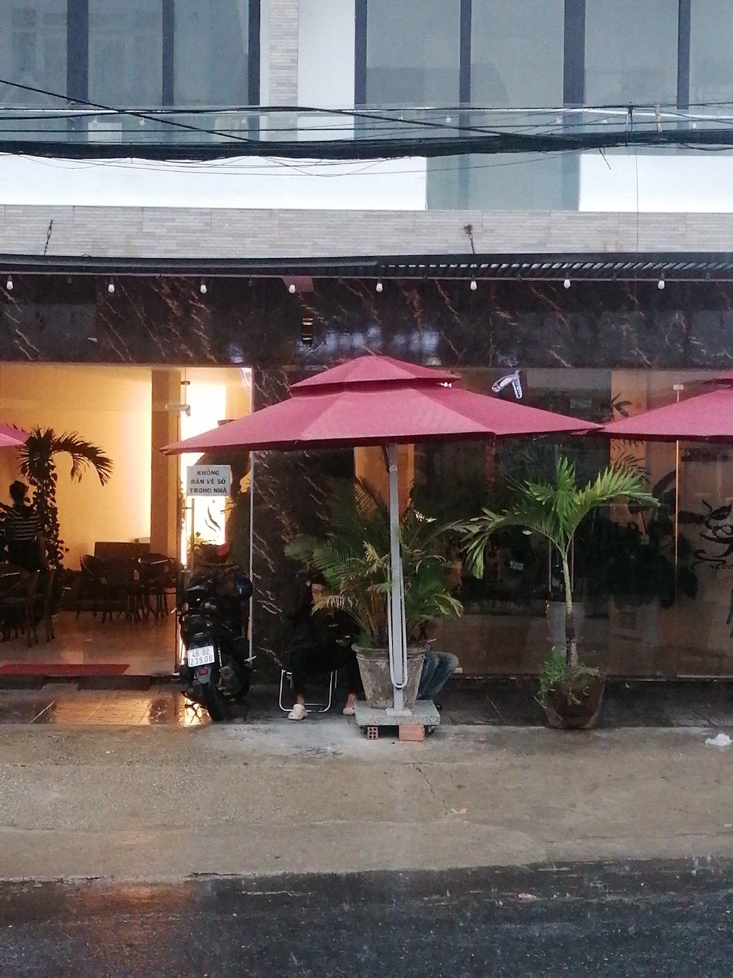 A street scene in Da Lat, Vietnam. The ground appears wet with rain. A building with a marble facade stands behind a row of red umbrellas. Under one of the umbrellas, two people are seated. A motorcycle is parked next to them. Inside the building, there is furniture and other people. A sign in Vietnamese is visible near the building's entrance. Overhead, electrical wires run along the facade.