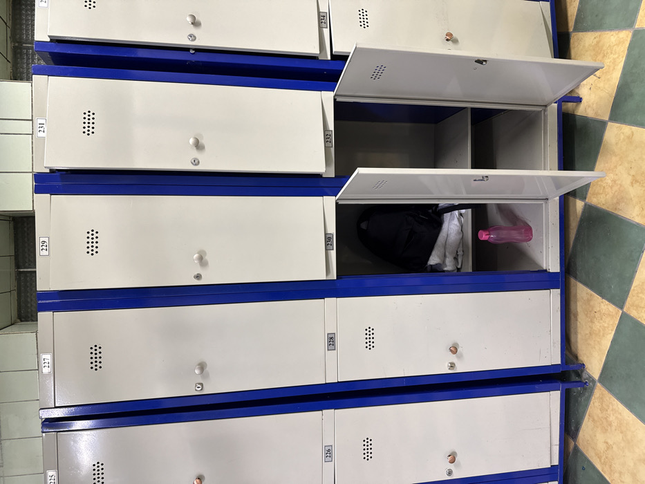 The image shows multiple rows and columns of metal lockers, typically found in a changing room, gym, or public facility. The lockers have white doors with a distinct blue frame or trim outlining each individual compartment. Each door features a round white knob and a keyhole below it, alongside a rectangular grid of small ventilation holes positioned on the upper left side of the door.
Small, rectangular white labels with black numbers are affixed to the vertical divisions between the lockers, indicating their unique identifiers. Visible numbers include 225, 226, 227, 228, 229, 230, 231, 232, 233, and 234. The numbering appears to proceed sequentially down vertical columns and then across to the next column.
Several lockers are open, revealing their contents or lack thereof. Locker number 230, located on the right side (second from the bottom row), is open and contains a black backpack or duffel bag, some folded white clothing (possibly a towel or t-shirt), and a translucent pink water bottle lying on its side. Locker 232, positioned directly above 230, is also open and empty, showcasing its blue interior and an internal horizontal shelf. Locker 234, partially visible at the top right, is likewise open and empty, also featuring an internal shelf.
The floor on the right side of the image is tiled with a checkerboard pattern of alternating light beige/brown and muted green/darker beige square tiles. On the far left, a wall covered with light-colored, rectangular tiles is visible. The lighting is even, suggesting an indoor environment, likely illuminated by artificial light. The perspective is slightly overhead, and the entire image is rotated, presenting the vertically stacked lockers horizontally across the frame.