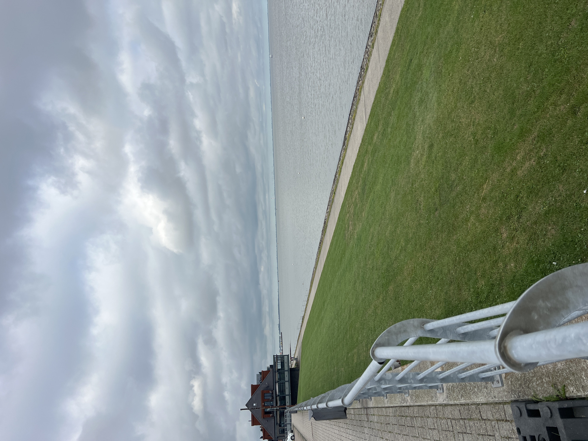 The image shows a view of the Jade Bay in Wilhelmshaven, Germany, from a paved walkway with a metal railing. The sky is overcast with thick, gray clouds. The water is calm and appears to be a pale gray color. On the left side of the image, there is a grassy embankment leading down to the water's edge. A building with a red roof is visible on the left, set back from the walkway. The building appears to be a restaurant or hotel. There are a few white buoys in the water and a small sailboat in the distance. The walkway is made of light-colored paving stones. The metal railing is silver and consists of several horizontal bars and vertical supports.