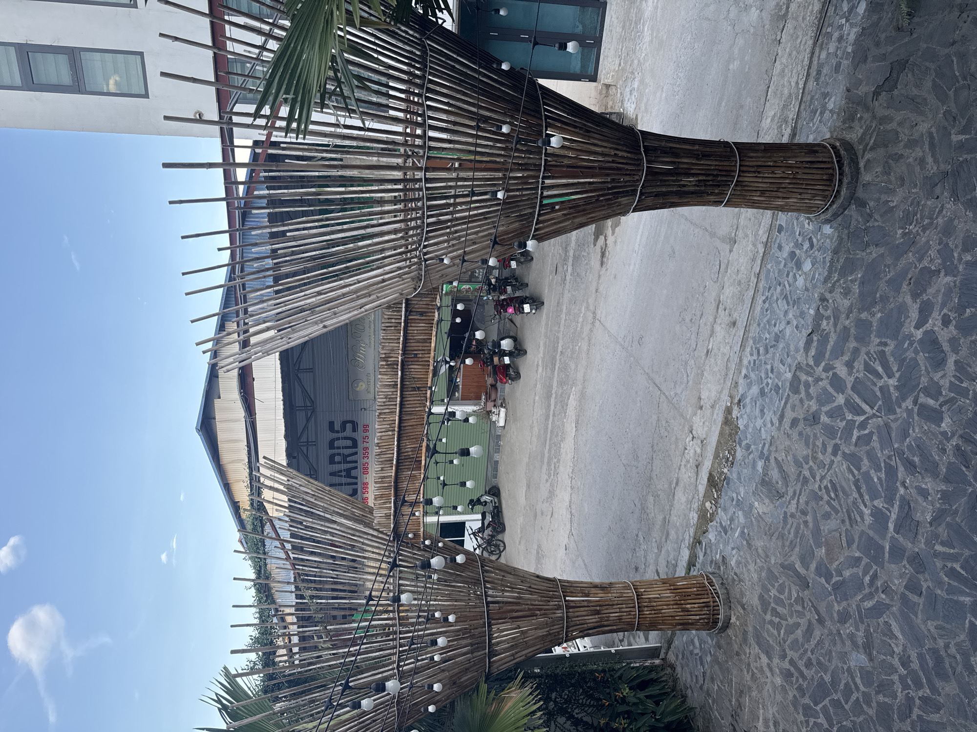 The image displays an outdoor scene in Trung Tam Ward, Vietnam, under clear daylight, with no individuals present. Two prominent, conical structures made of bundled bamboo poles serve as decorative pillars, framing an entrance in the foreground. Strung between these bamboo pillars and extending across the midground are numerous string lights with white bulbs. The immediate ground is paved with irregularly shaped dark gray flagstones, transitioning to a lighter gray concrete surface beyond. Parked on this concrete surface are several motorbikes and one bicycle. In the mid-background, a green building or wall is visible, also featuring string lights. The background includes a large gray building with a sign displaying "LIARDS" (likely "Billiards") and a phone number: "36 598 - 085 359 75 99". Adjacent to this, a taller, white multi-story building with dark rectangular windows is visible. The sky is clear blue with a few scattered white clouds, and green foliage is present on the left and right edges of the frame.