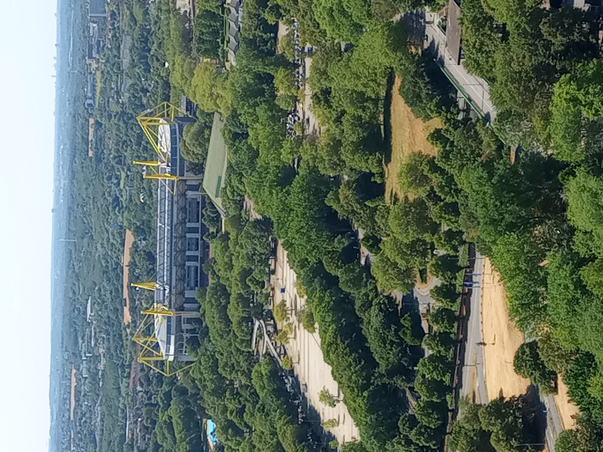 This is a high-angle aerial view of the Signal Iduna Park stadium in Dortmund, Germany, on a clear, sunny day. The stadium, iconic for its yellow pylons, is surrounded by a dense urban forest. In the foreground, a large grassy field with some dry patches is visible, bordered by trees and a road. Further back, a large parking lot with a few cars is situated amidst the trees. A blue swimming pool with a surrounding deck can be seen to the left of the stadium. The cityscape stretches out into the distance, with buildings and more greenery visible under a hazy sky. The overall impression is a large, modern stadium integrated into a lush, green urban environment.