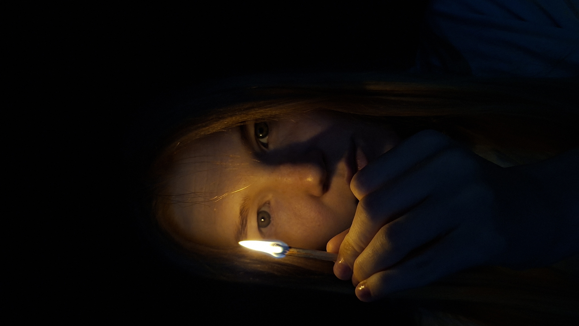 A close-up, waist-up shot captures a young person with fair skin and auburn hair holding a lit match in their right hand in front of their face against a dark background. The flame illuminates the person's face from the left, casting shadows and highlighting their facial features, including freckles. Their eyes, which appear to be green or hazel, are looking directly at the viewer. The hand holding the match is positioned near the lower center of the frame, with the fingers slightly curled. The overall lighting is dim, with the flame being the primary light source, creating a dramatic and intimate mood. The image does not contain any identifiable elements that would suggest a specific location within the city of Большой Букор, Russia.