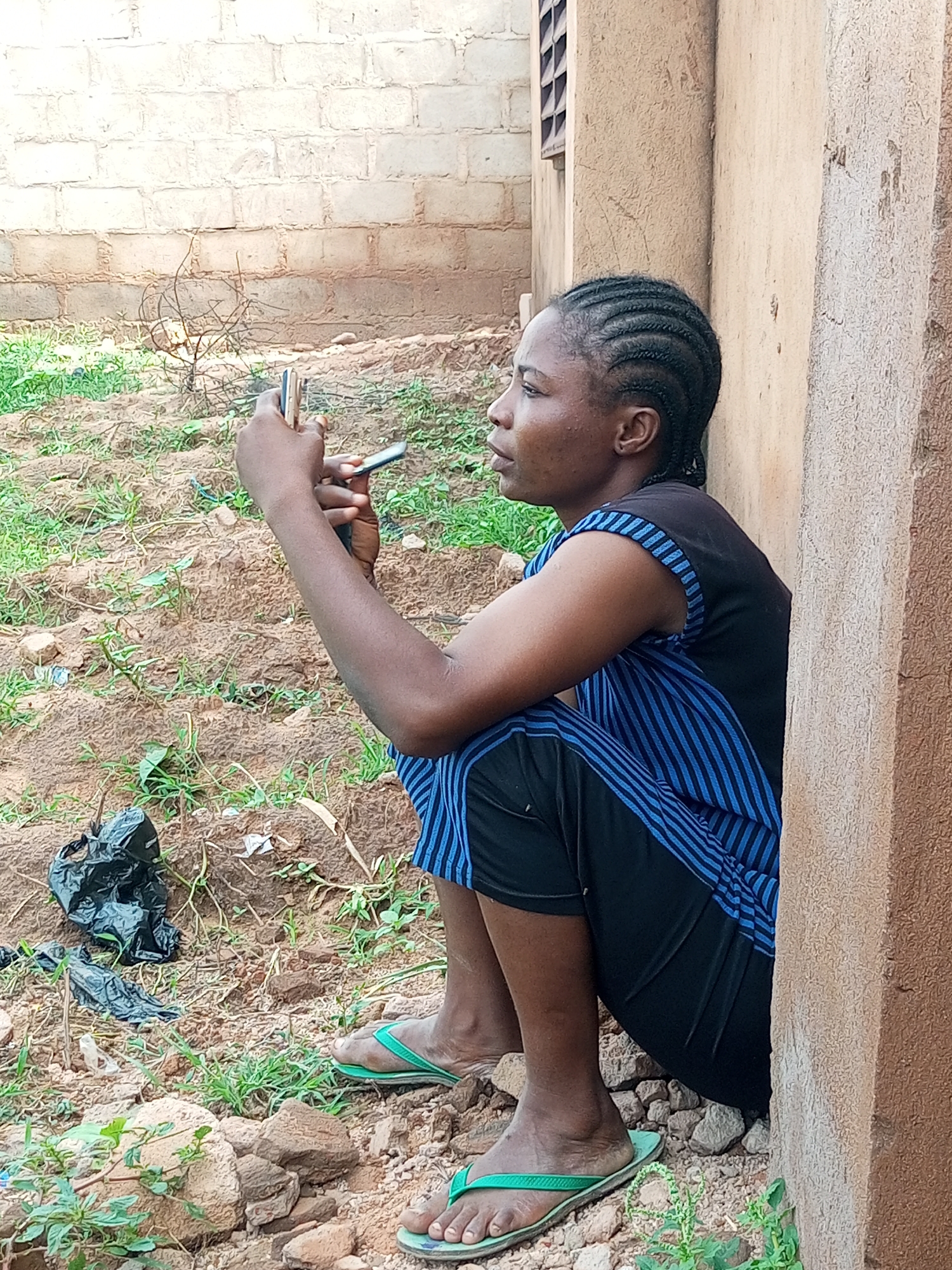 A young Black woman with braided hair sits against a rough, textured, light brown wall in Tempure, Nigeria. She is wearing a black sleeveless top with blue stripes around the armholes and black pants with blue stripes down the sides. She is holding two mobile phones in her hands. She is sitting on rocky, dry ground with sparse green vegetation. To her left is a black plastic bag on the ground. The background consists of a light-colored brick wall.
