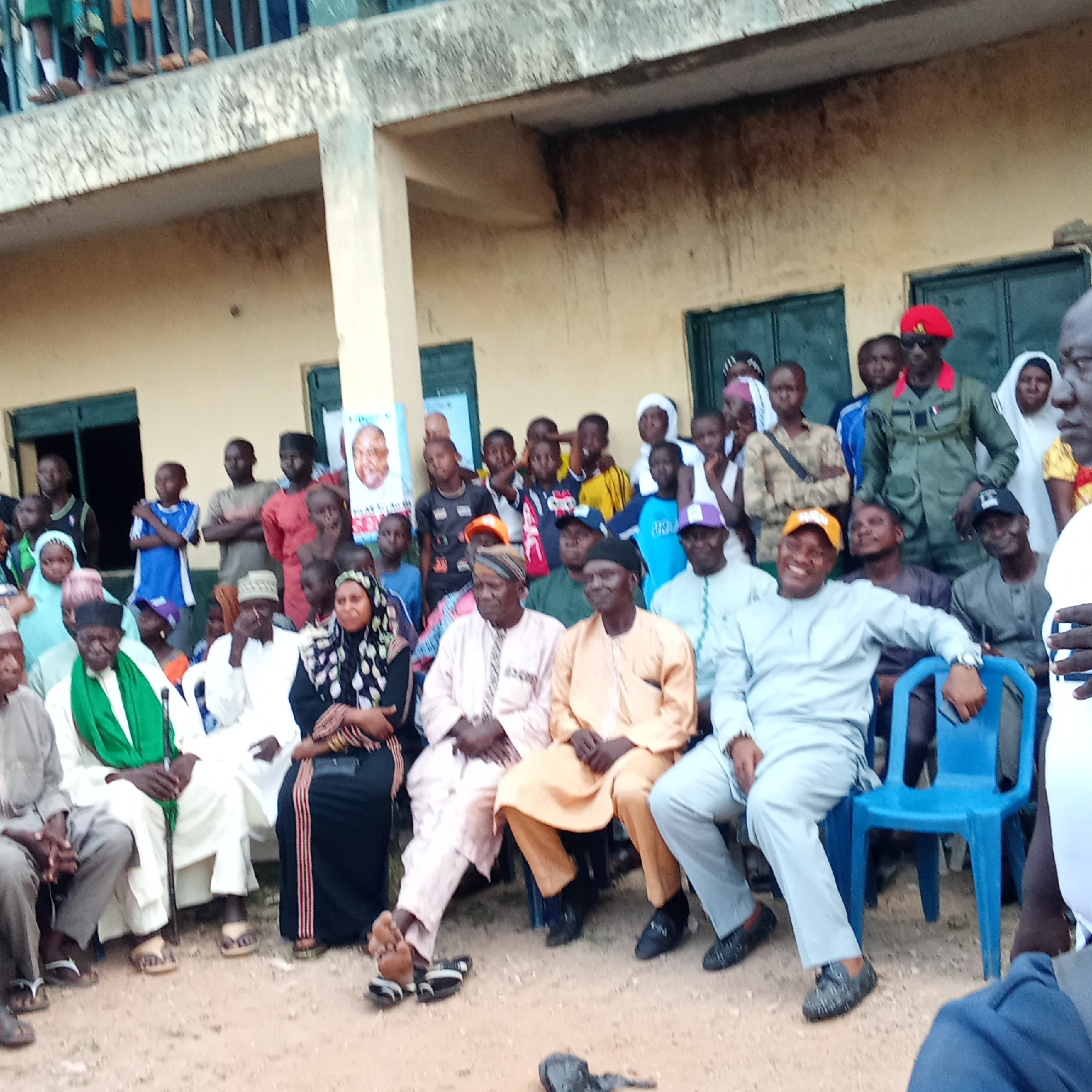 A large group of people are gathered outdoors in Akwanga, Nigeria. Most are seated, a few are standing. The group consists of men, women, and children of varying ages. Many of the men are wearing traditional attire, including robes and hats. One man standing wears a military-style uniform with a red beret. Some women wear hijabs. A poster with a man's face is affixed to the building behind the group. A blue plastic chair is visible. The building appears to be a single-story structure with green doors and windows. Some people are standing on an elevated outdoor platform, possibly a balcony. The ground is dirt.