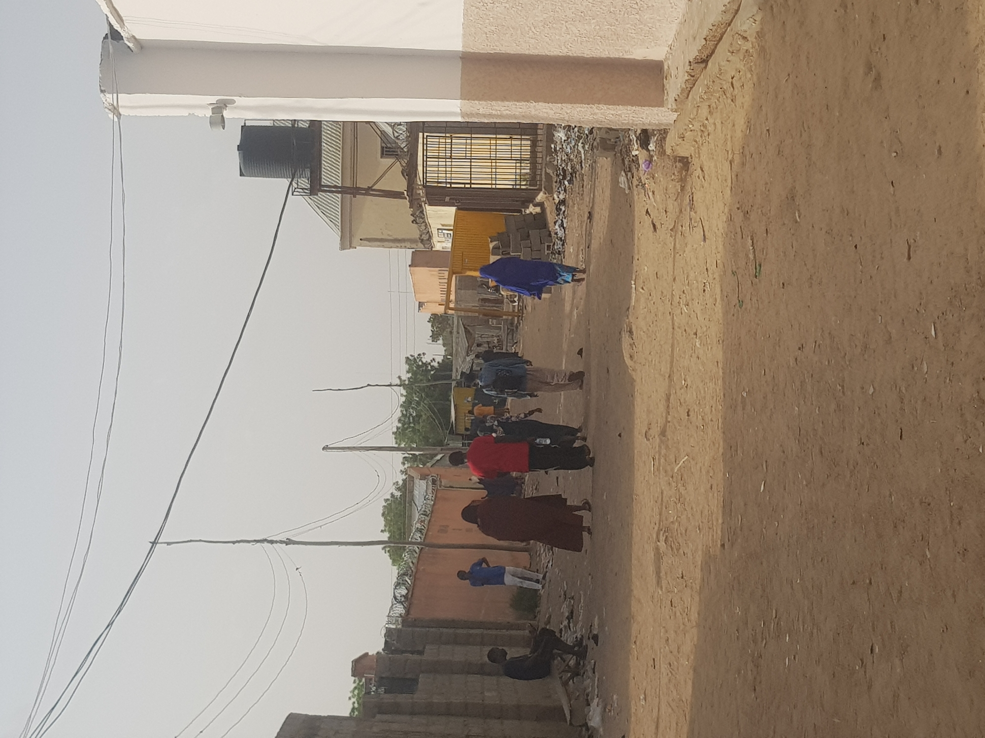 The image shows a street scene in Dabawa, Nigeria. The street is unpaved and sandy. Several people are walking along the street. One person is wearing a blue garment, another a red shirt, and yet another a brown robe. Some people are standing near a partially constructed building made of concrete blocks. Electrical wires are visible overhead. Buildings line the street, varying in construction quality. One building has a water tank on its roof. There is some debris on the sides of the street. The sky is overcast.