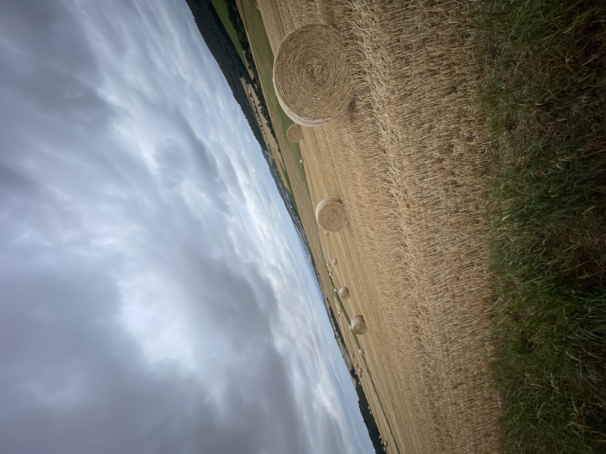 The image depicts a rural landscape with a field of harvested wheat, evidenced by numerous cylindrical bales of straw scattered across the golden stubble. The field slopes gently downwards from right to left, with a dense, dark green forest bordering the middle ground on the left. In the distance, a town or village is visible, nestled at the foot of rolling hills that rise to the left. The sky is heavily overcast with dark, dramatic clouds, suggesting an impending storm. The overall scene conveys a sense of rural tranquility disrupted by the approaching weather. There are no people visible in the image. The image's perspective is from a slightly elevated vantage point, looking across the field. The presence of hay bales indicates agricultural activity. The scene is situated in or around Asterode, Germany, a location known for its agricultural lands and forested hills.