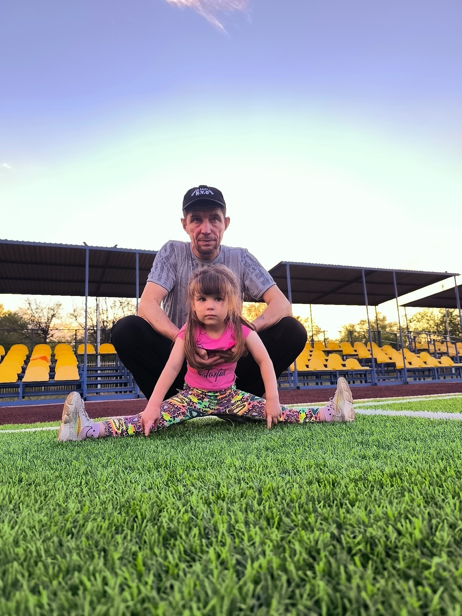 A low-angle shot captures a man and a young girl on a green turf field, likely a sports stadium, with rows of yellow stadium seats in the background. The man, wearing a grey t-shirt and a black baseball cap, is crouched behind the girl. The girl, dressed in a pink top and colorful leggings, is performing a split with her legs extended in opposite directions. Both are looking towards the camera, with the girl's hands resting on her knees. The sky above is a gradient of blue and light yellow, suggesting it is either sunrise or sunset. The location is not specified in the city of Qarağandı, Kazakhstan.