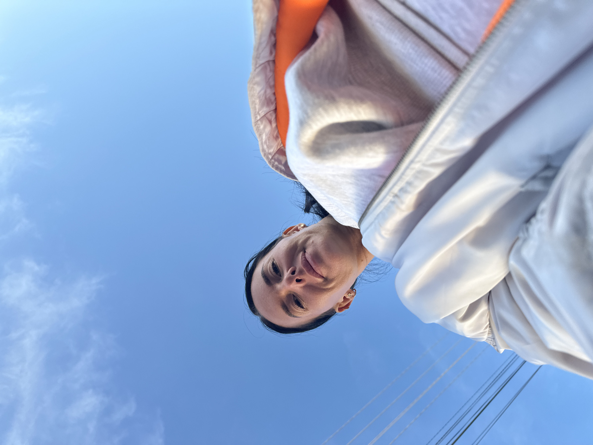 This image is a low-angle shot looking up at a woman against a clear blue sky, with the location context being Ольховка, Russia. The woman's face is centered in the lower portion of the frame, as she looks directly down at the camera with a neutral, slightly smiling expression. She has dark hair and wears small gold hoop earrings. She is dressed in a light-colored jacket, possibly white or light grey, which features a distinct orange lining around the collar and hood area. The vast majority of the background consists of a bright, uninterrupted blue sky, with only a few faint, wispy white clouds visible in the upper left. In the lower right of the image, several dark power lines run diagonally across the frame, suggesting an outdoor, possibly developed, setting. The clear sky and bright illumination indicate that the photo was taken during daytime with good weather. There is no visible text in the image. The unique perspective implies a selfie or a photograph taken from directly below the subject.