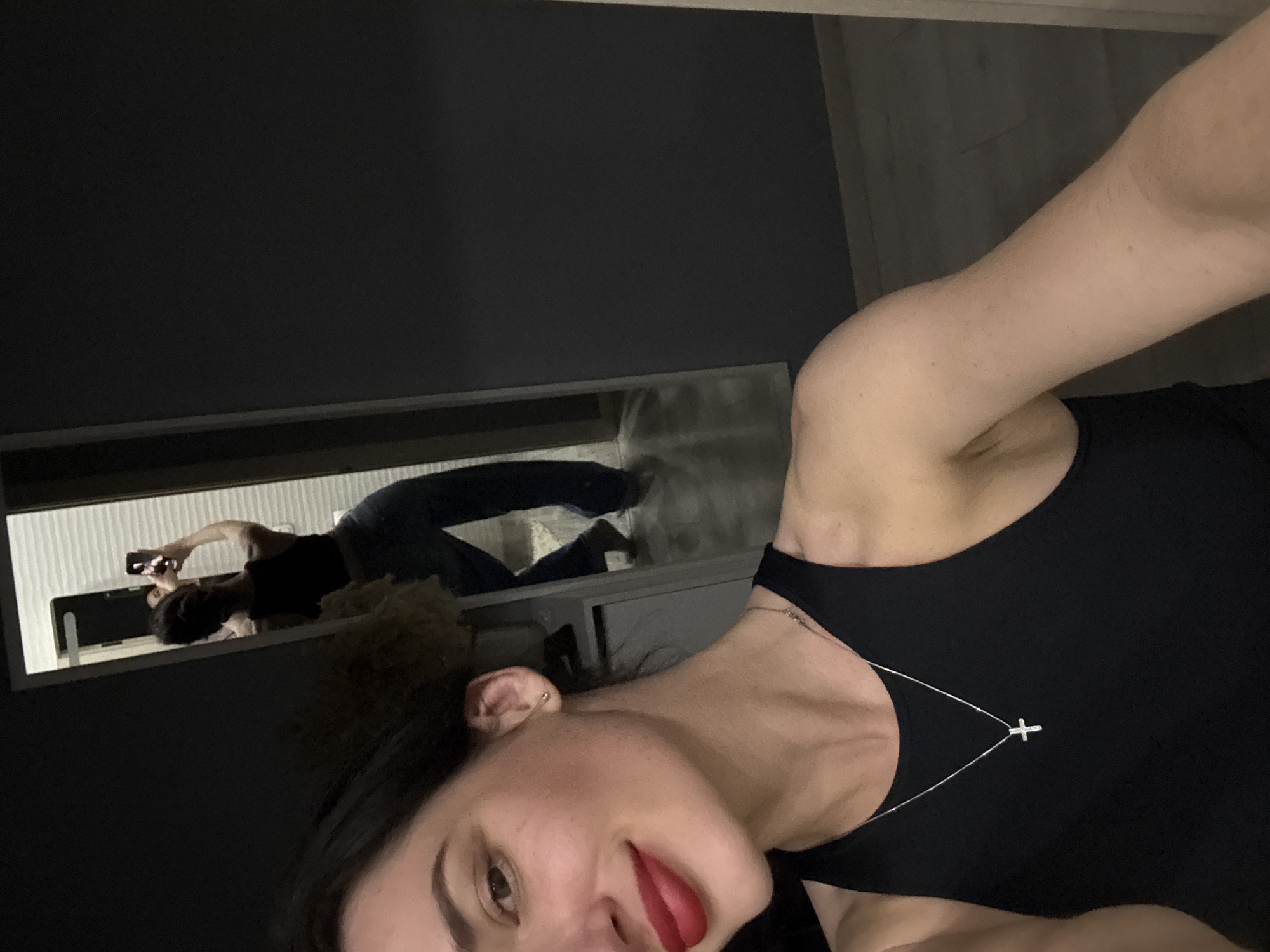 A selfie of a woman wearing a black top and a silver necklace with a cross pendant. She is looking at the camera with red lipstick and a dark hairband. In the background, a mirror reflects the woman taking the selfie, wearing jeans and a black top, holding a phone in her hand. The walls are dark, and the location is Unknown, Unknown.