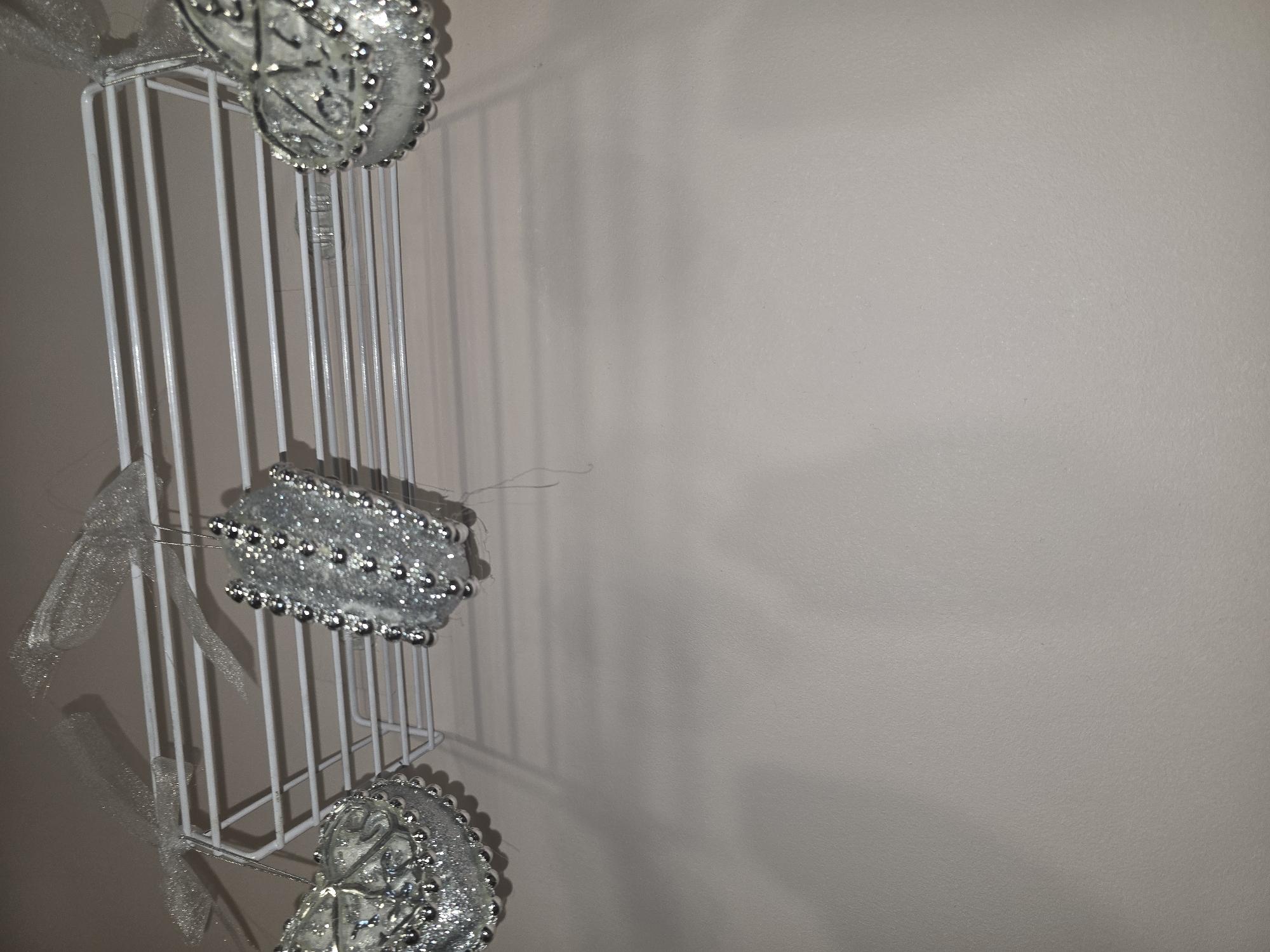 A white wire shelf is mounted horizontally on a wall. Three heart-shaped ornaments are hanging from the shelf by thin strings. The ornaments are silver and decorated with small beads and glitter. One ornament is on the left, another in the center, and the third on the right. A light-colored wall serves as the background. There is no indication of the location being in Ulan-Ude, Russia, or any other specific city.