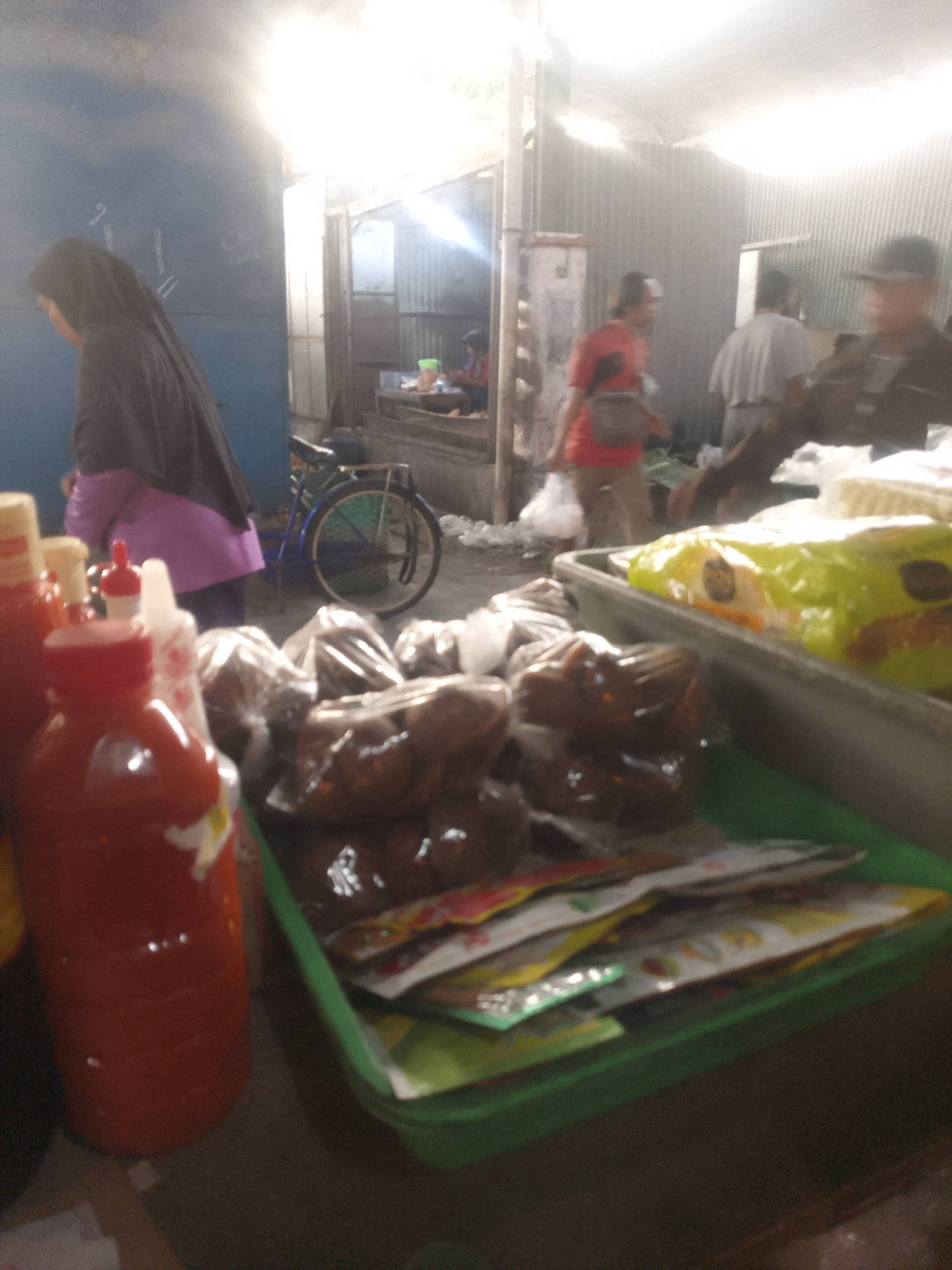 This is a close-up, slightly blurry, indoor photograph of a market stall in Gondangrejo, Indonesia. In the foreground, a bright red bottle of chili sauce or a similar condiment stands prominently on a dark surface. To the right of the sauce bottle, a green plastic container is filled with various small, individually packaged food items, possibly snacks or spice mixes, layered on top of each other. Some of these packages have colorful designs. Behind the green container, a large, bright yellow plastic bag of food products is visible. In the background, there are people in the market. To the left, a person wearing a dark hijab and a purple garment stands near a blue wall. Further back, several individuals are standing or walking, some appearing to be vendors or customers. One person in the middle ground wears a red shirt and appears to be carrying a bag. Another person is visible to the right, possibly wearing a hat. The environment suggests an open-air or covered market with corrugated metal walls visible in the distance. The lighting is bright and diffused, with a strong light source coming from the upper right of the frame, possibly indicating overhead lighting or daylight.