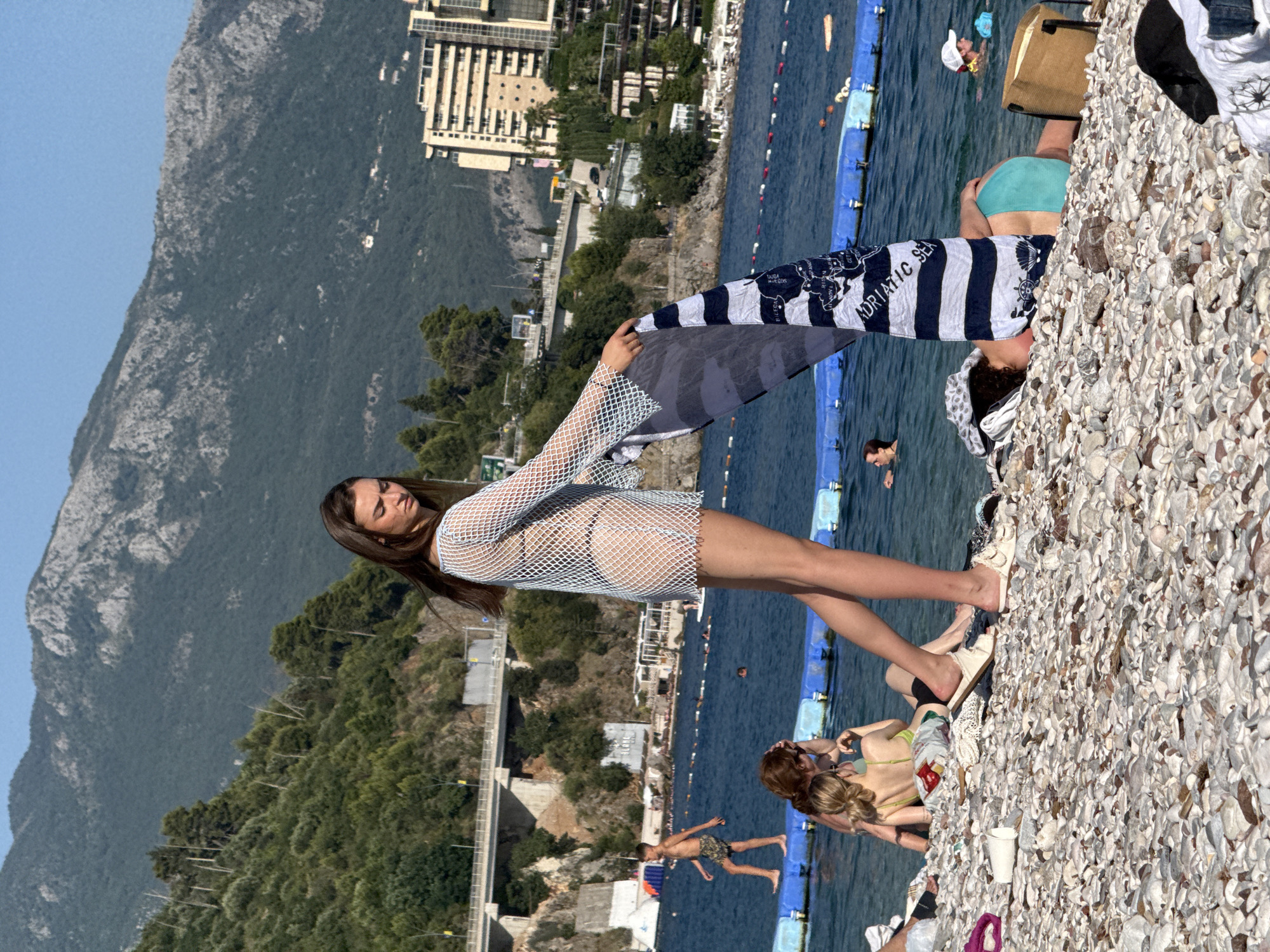 In Budva, Montenegro, on a sunny day at Mogren Beach, a young woman stands on a pebble shore, holding a striped beach towel that reads "Adriatic Sea." She wears a white mesh cover-up over a bikini. In the background, the blue Adriatic Sea is dotted with swimmers and inflatable barriers. Across the water, a coastal road with vehicles and a bridge winds along a forested hillside. Beyond the hill, modern buildings rise against a backdrop of imposing, rugged mountains. On the beach, other beachgoers relax and swim.