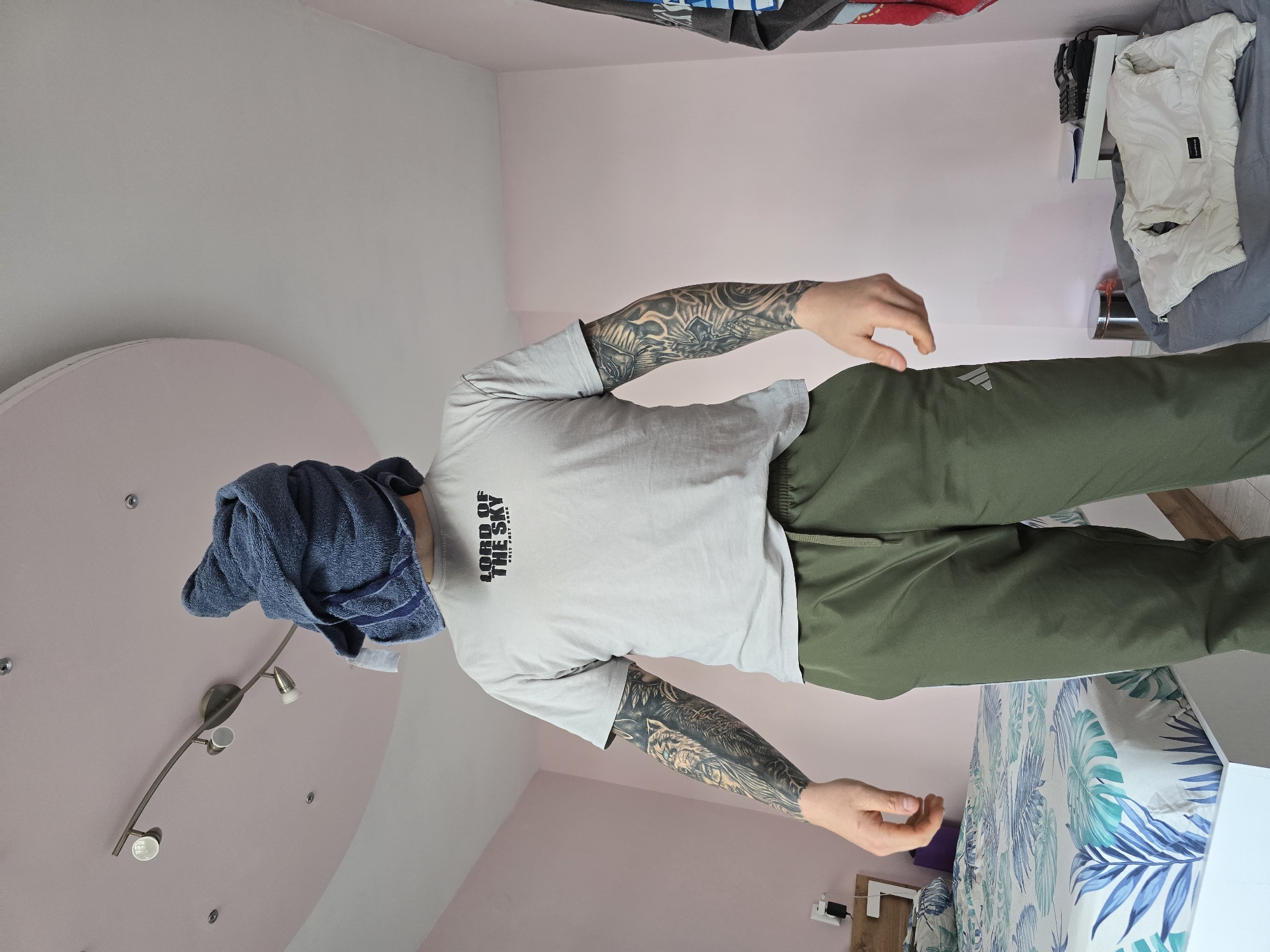A man stands indoors with a blue towel over his head. He wears a gray t-shirt with the words "LORD OF THE SKY" on it, and green Adidas pants. The man has extensive tattoo work on both arms. The room has pink walls. A bed with a blue and green tropical leaf patterned comforter is visible. There is a light fixture on the ceiling. The location is inferred to be Burgas, Bulgaria, only due to the prompt's explicit instructions to include that location.