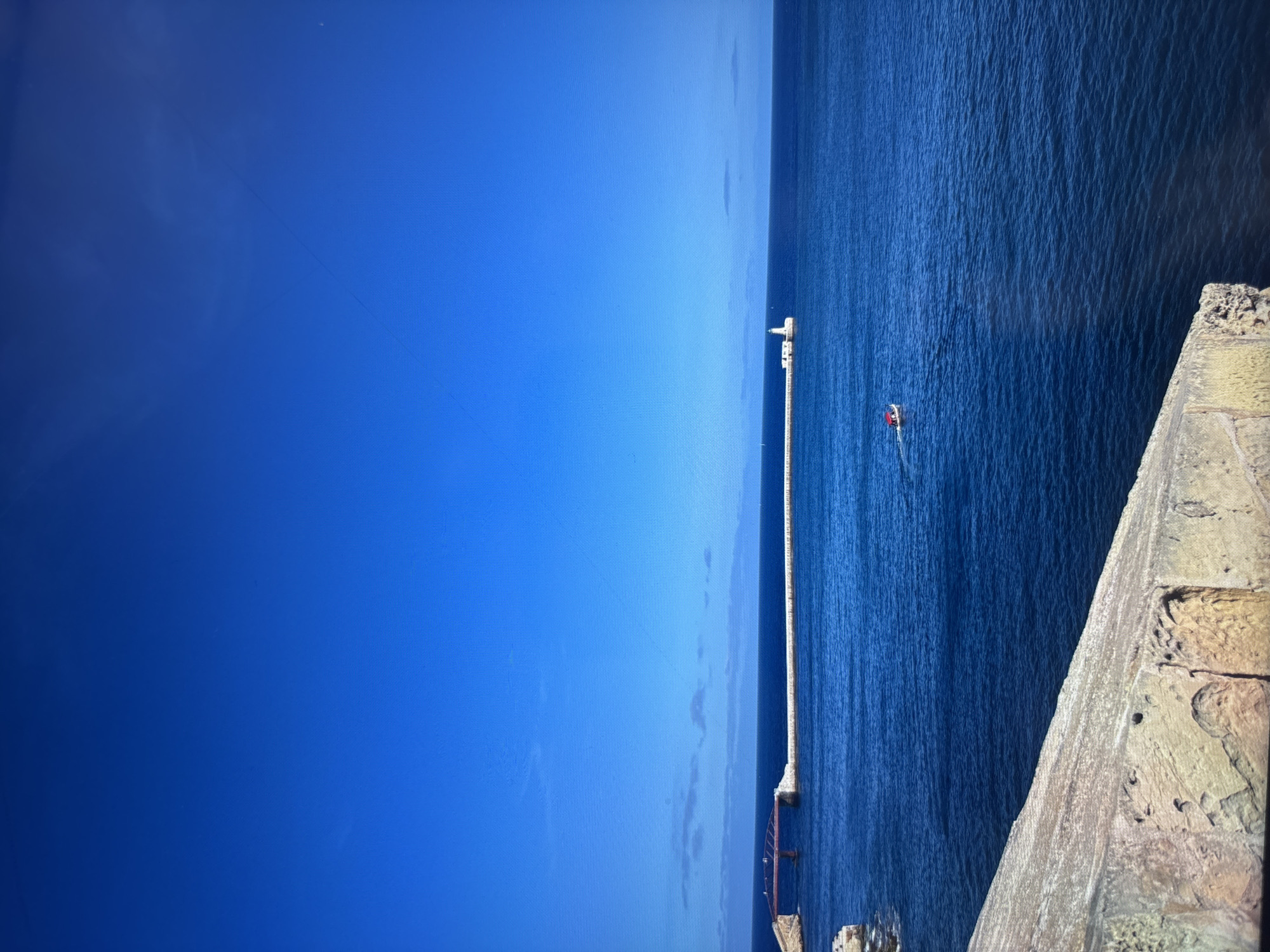 The image depicts a view of the sea and sky, seemingly taken from a high vantage point. A long, light-colored pier stretches out into the water. A red object, possibly a boat or buoy, is visible near the pier. A stone structure is located in the lower right corner of the image, suggesting the viewpoint is from a building or wall. The location appears to be a coastal area, but the specific city is not identifiable from the visual information provided.