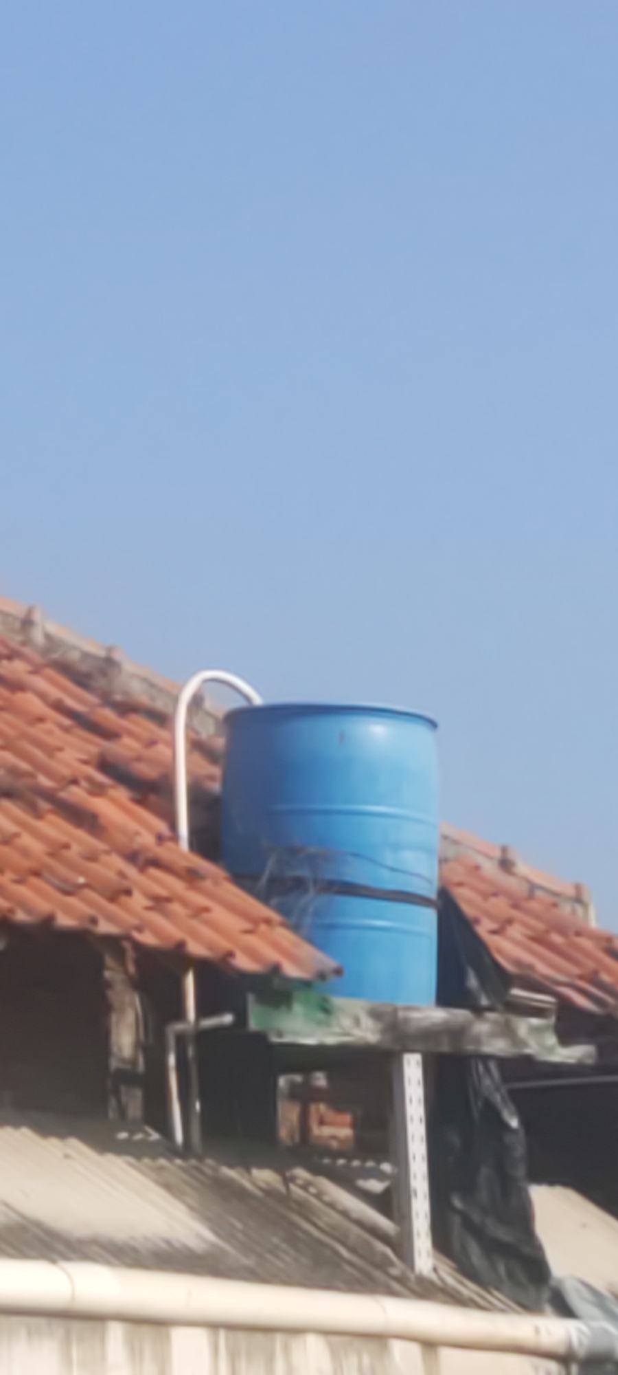The image shows a blue water barrel on a rooftop in Gebang, Indonesia. The barrel is cylindrical and has a white pipe connected to it. The rooftop is covered with orange tiles and appears to be part of a residential building. The sky is clear and blue. The image is taken from a low angle, looking up at the barrel and rooftop.