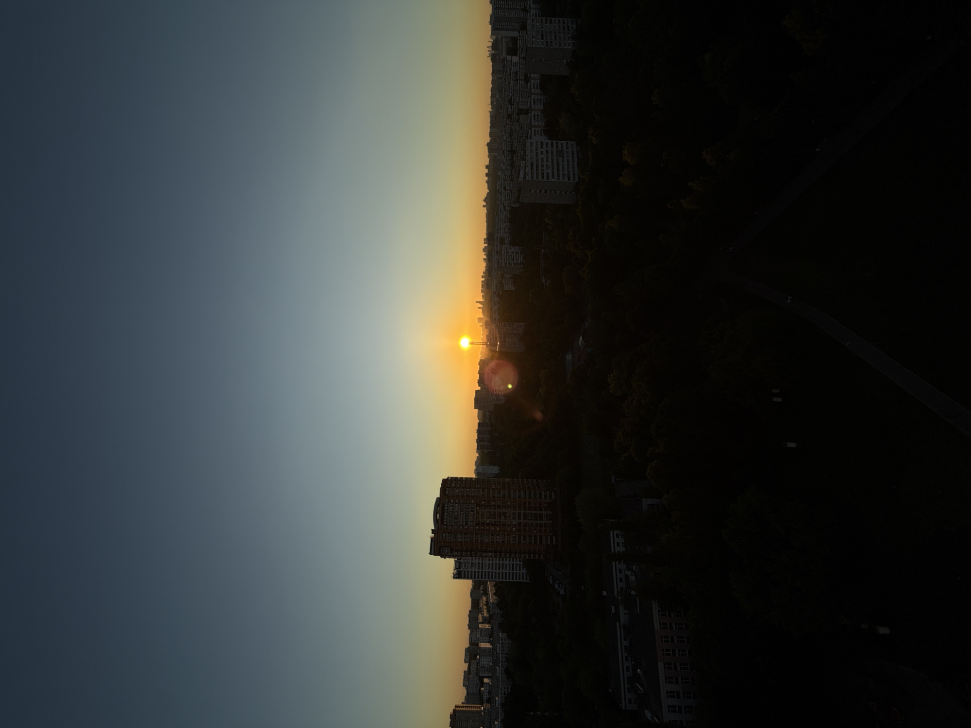 The image depicts a panoramic urban landscape at either sunrise or sunset, characterized by a clear sky transitioning from a deep indigo-blue overhead to a warm orange and yellow hue near the horizon. The sun, a brilliant point of light, is positioned low on the horizon slightly to the right of center, casting an intense glow and producing a circular lens flare in the lower right quadrant of its vicinity.
In the midground, a prominent, multi-story reddish-brown residential tower stands out against the sky, flanked by a taller, slender white building to its left. Numerous grey, multi-story apartment buildings, consistent with Soviet-era architectural styles, stretch across the horizon, forming a dense urban sprawl. Interspersed among these structures are extensive areas of dark green trees, forming a significant canopy.
The foreground is enveloped in deep shadow, rendering details largely indistinct. Visible elements include a mass of dark trees, a shadowy, low-rise building with faintly visible window patterns on the far left, and a curved, dark asphalt path or road winding through the lower right. No specific human figures or discernible activities are visible across the scene.
The architectural styles, particularly the prevalence of the grey apartment blocks and the dense integration of green spaces, are highly characteristic of a residential district within the city of Moscow, Russia. The low sun suggests the camera is facing either east (sunrise) or west (sunset).