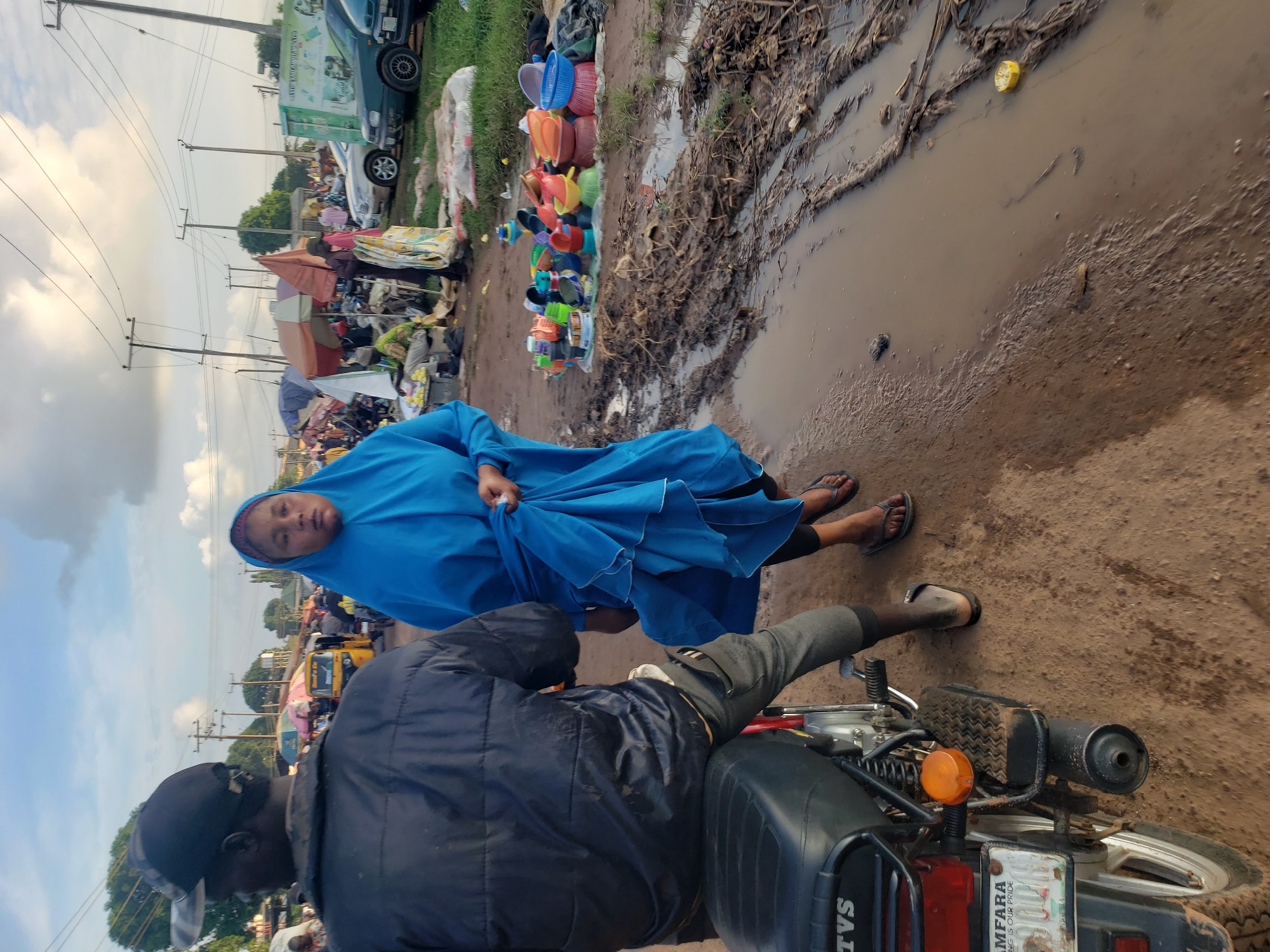 In Kakuri Gwari, Nigeria, a woman wearing a blue hijab and a flowing blue dress walks on a muddy road next to a motorcycle. She is holding her dress with her left hand. The road is covered in puddles and debris. In the background, a market is visible with stalls and several people. The sky is partly cloudy.