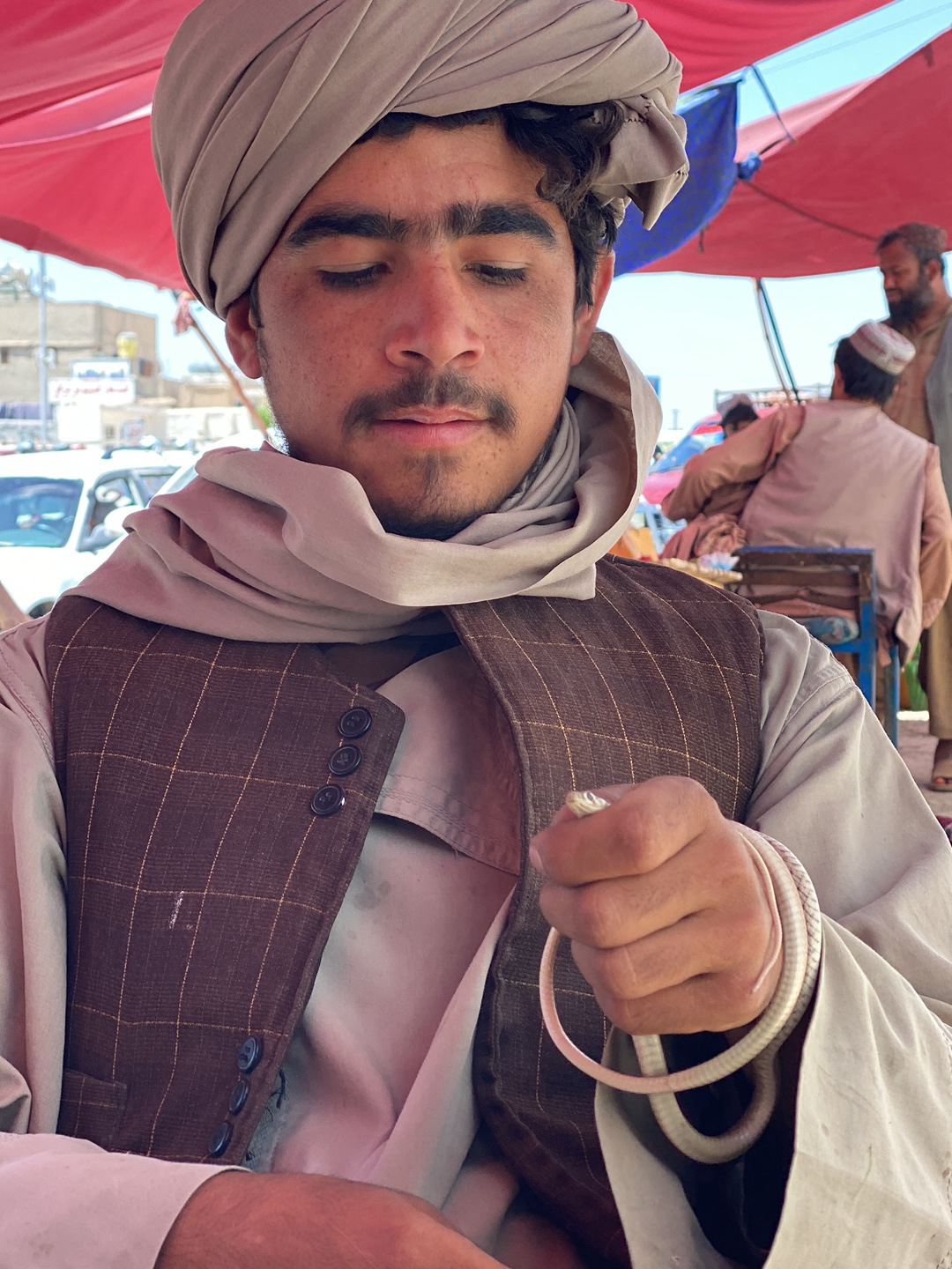 A man is seated in what appears to be an outdoor market setting in قندهار, Afghanistan. He is wearing a light-colored turban and a brown patterned vest over a tan or light-colored shirt. The man holds a small, light-colored snake coiled in his right hand, and he is looking down at it. In the background, people are sitting and standing around tables. There are vehicles and buildings visible in the far background. The environment suggests a busy, urban setting during daylight hours.