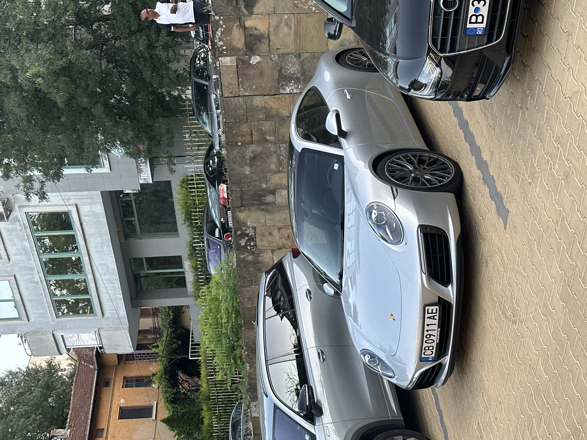 A silver Porsche 911 Carrera is parked on a light brown brick paver surface. The car's license plate reads "CB 0911 AE". To the right of the Porsche is the front end of a black Audi A4, also parked. Behind the Porsche and to the left is a silver SUV. Further back and uphill, a man in a white polo shirt and dark pants is standing and looking at his phone. The scene is in Veliko Tarnovo, Bulgaria, in front of a building with a light gray facade and green-tinted windows. To the left of the modern building is an older, yellowish building with a red tile roof. Trees and greenery are visible throughout the image, with a stone retaining wall behind the parked cars.