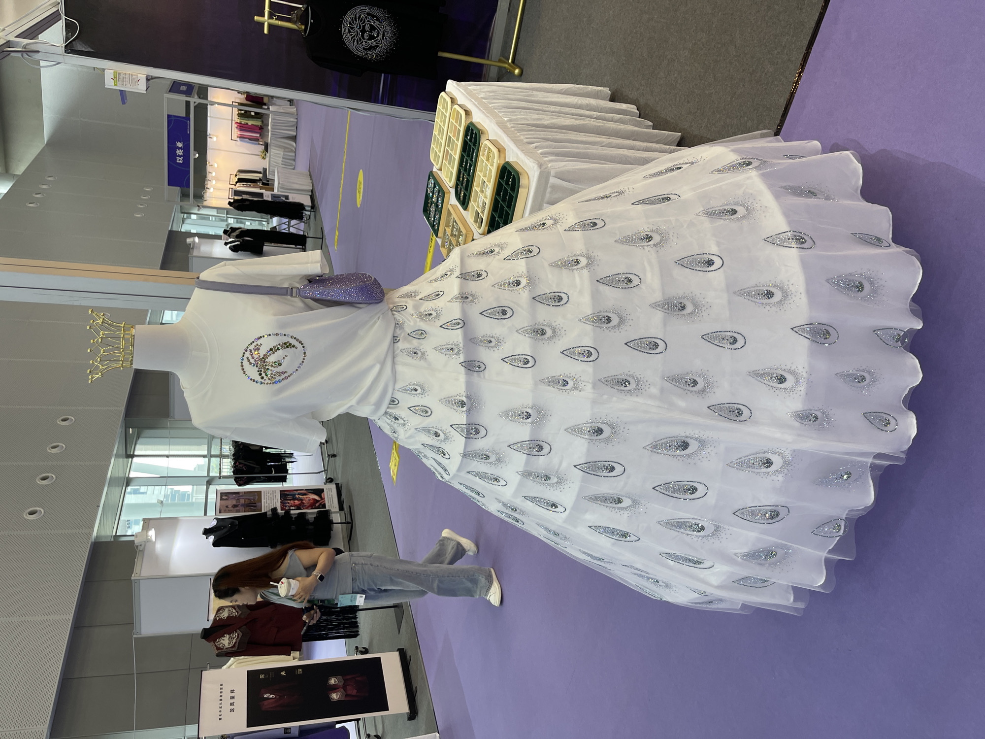 An exhibition booth in Haizhu District, China features a white mannequin wearing a white t-shirt with a sparkling emblem and a full, white skirt decorated with shimmering, teardrop-shaped appliques. A small, purple, beaded handbag hangs from the mannequin's shoulder, and a gold crown sits atop its head. Adjacent to the mannequin, a table draped with a white tablecloth displays several trays of jewelry. In the background, a woman wearing a grey off-the-shoulder top and blue jeans holds a cup and a phone. Several other exhibition booths and clothing racks are visible, with signs in Chinese. The flooring is purple carpet.