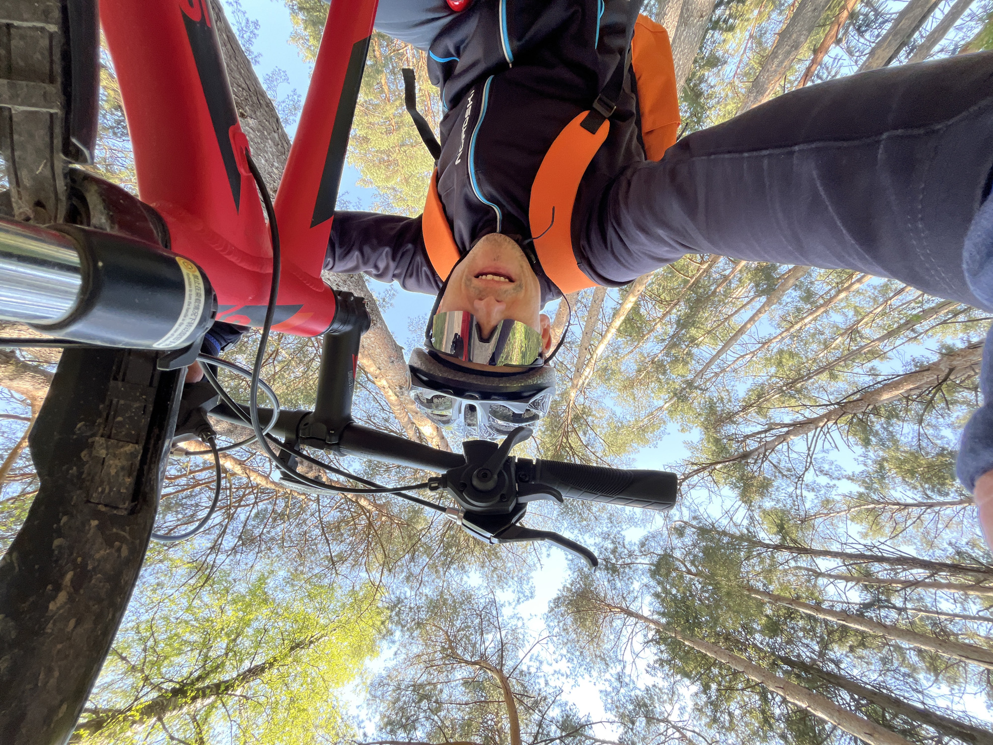 A low-angle, perspective-distorted image shows a man on a red mountain bike in a wooded area. The man is wearing a helmet and sunglasses, with a dark jacket and an orange backpack. The view is looking upward, capturing the sky and treetops. The location is likely a forest or park near Chaikovsky, Russia, based on the prevalence of pine trees. The man is looking down into the camera, suggesting he took a selfie while on the bike.