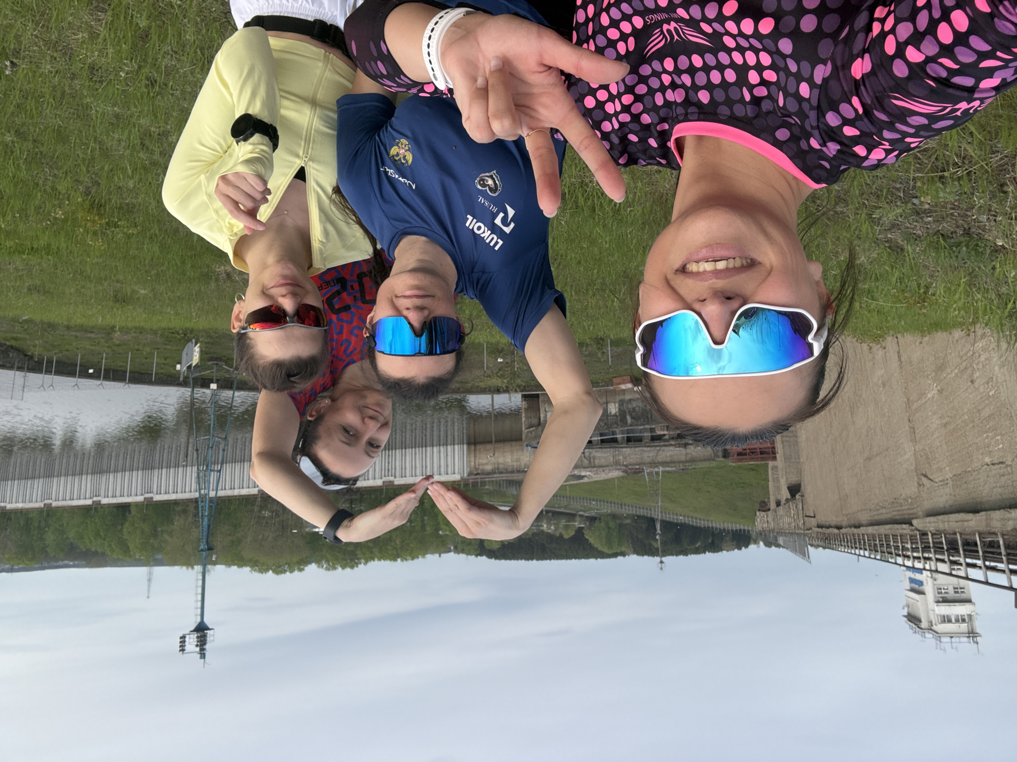 Three women are posing outdoors near a dam, with the cityscape reflected in their sunglasses. The woman on the right wears a pink and purple polka dot athletic shirt and makes a peace sign. The woman in the center wears a blue athletic shirt with the logo "LUKOIL." The woman on the left wears a yellow jacket. The location is likely the city of Chaikovsky, Russia, based on the Russian logos and Cyrillic text on the blue shirt. The women have their arms extended and hands forming a heart shape with their fingers, reflecting the skyline.