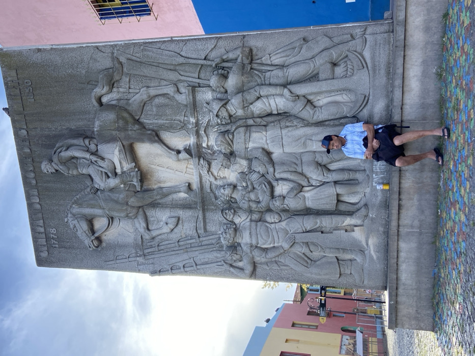 A horizontal shot shows a gray stone relief of a battle scene with a man posing beside it. The relief features figures on horseback and soldiers. In the bottom right, a man in a light blue shirt, black shorts, and a dark blue hat stands with his arms crossed. Behind the relief, the sky is visible along with colorful buildings and an overcast sky. The ground appears to be paved with colorful tiles.