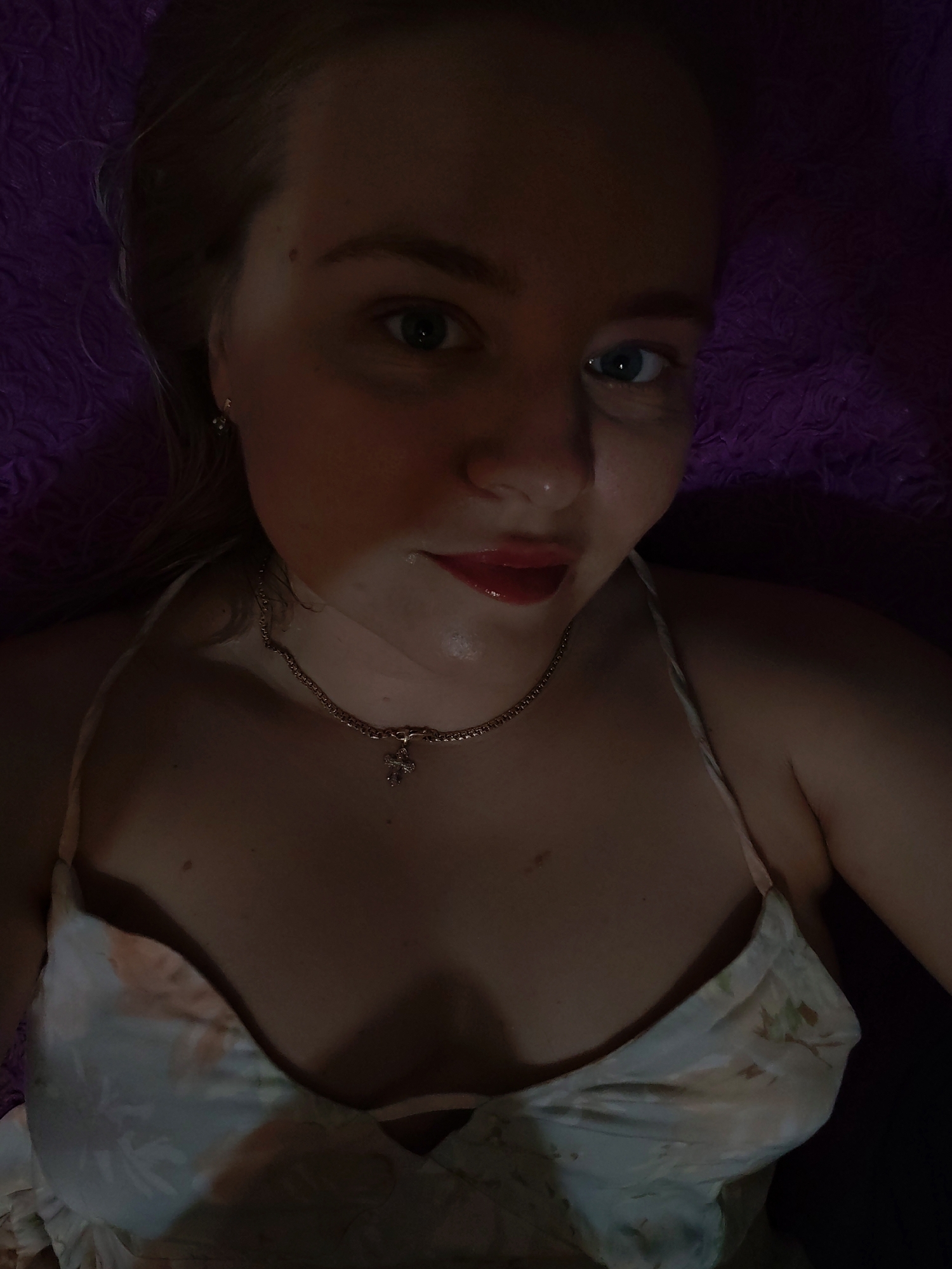 A woman with fair skin and blond hair is lying on a purple surface. She is wearing a floral-patterned top with thin straps and a gold chain necklace with a cross pendant. Her lips are glossy and slightly parted. The lighting in the image is dim, creating shadows on her face and body. The location is unknown and cannot be determined from the image.