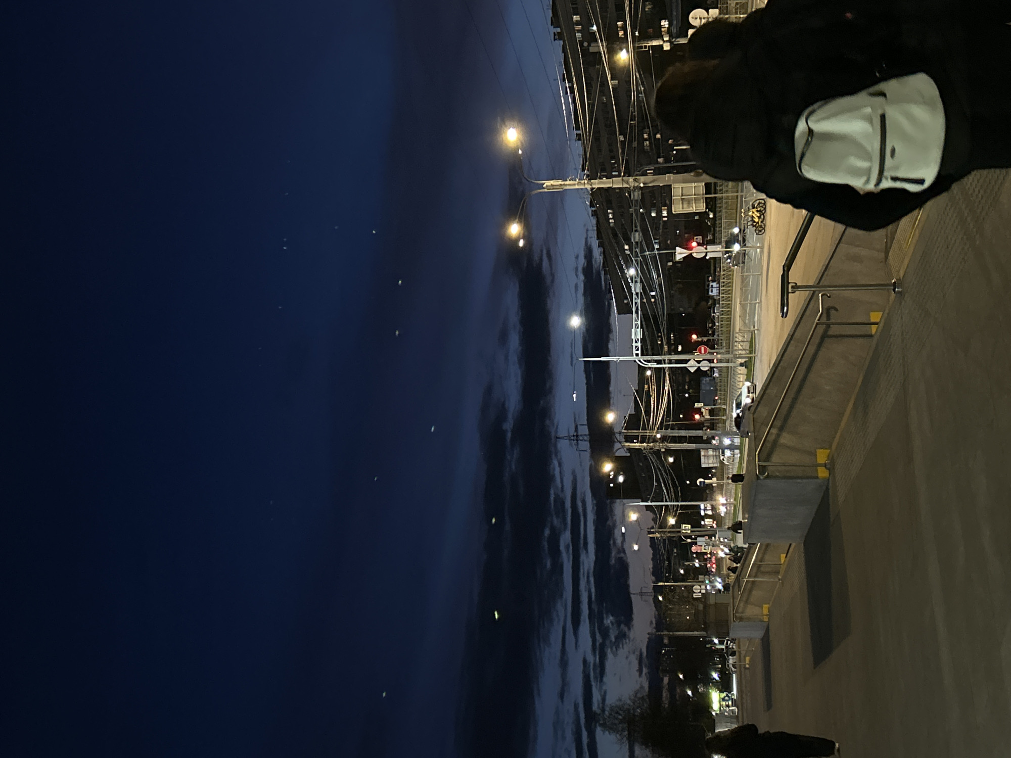 An outdoor, urban night scene is captured from an elevated perspective, rotated ninety degrees counter-clockwise. When viewed upright, a dark blue-black sky with scattered clouds occupies the top. Several small, bright, blurry white and yellow light sources are visible against the dark sky.
Below the sky, the midground reveals an intricate network of overhead wires and utility poles, characteristic of an electrified transport system like trams or trains. Numerous streetlights emit warm yellow and orange glows, illuminating the surroundings and creating some lens flares. Distant buildings show illuminated windows against the darkness.
On the right side of the midground, a street level is visible with traffic lights (red and green) and road signs. A bicycle rack containing several bicycles is discernible near the traffic lights. Blurry figures of people can be seen on the street or an adjacent platform.
In the foreground, on the bottom right, a concrete or paved platform with a metal handrail is prominent. A person, viewed from behind, stands on this platform, wearing a dark jacket and a white shoulder bag or backpack, appearing to observe the scene below. The overall impression is a bustling urban environment at night in Saint Petersburg, Russia. No discernible text is visible.