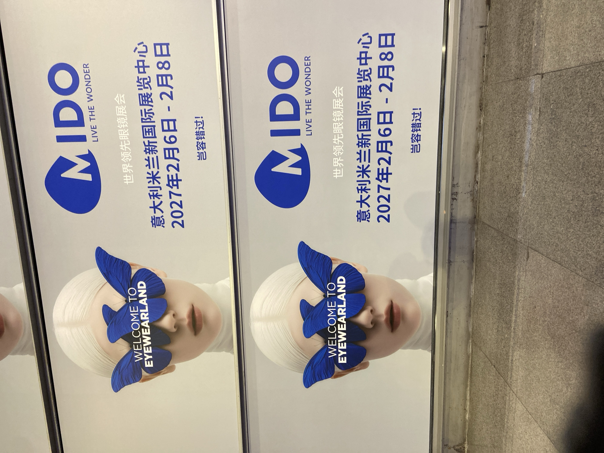 The image displays two identical billboards for the MIDO eyewear exhibition. Each billboard features a close-up of a person's face with white hair, wearing a white turtleneck. Blue butterfly-shaped eye coverings with the text "WELCOME TO EYEWEARLAND" are placed over their eyes. The MIDO logo and event details, including "LIVE THE WONDER," "世界领先眼镜展会" (World Leading Eyewear Exhibition), "意大利米兰国际展览中心 2027年2月6日 - 2月8日" (Fiera Milano International Exhibition Center, February 6-8, 2027), and "岂容错过!" (Don't miss it!), are present in Chinese. The billboards are positioned horizontally, one above the other, in what appears to be an outdoor urban environment. The lower portion of the image shows a tiled ground surface and a dirty, weathered wall. While the location is identified as Futian District, China, there are no discernible landmarks to confirm this.